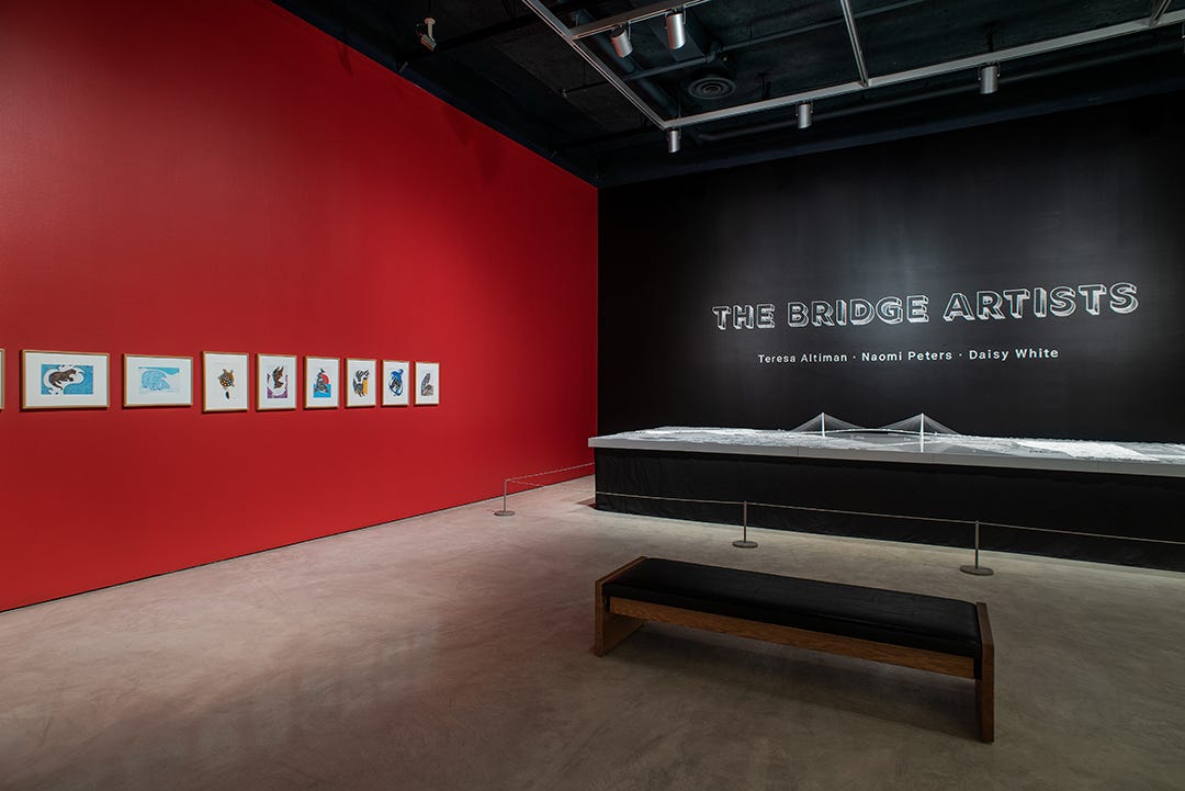 To the left, eight of Naomi’s drawings from the Indigenous Animal Series are in view against a dark red wall. To the right of the drawings is a dark black wall that says “The Bridge Artists” in capitalized white font.