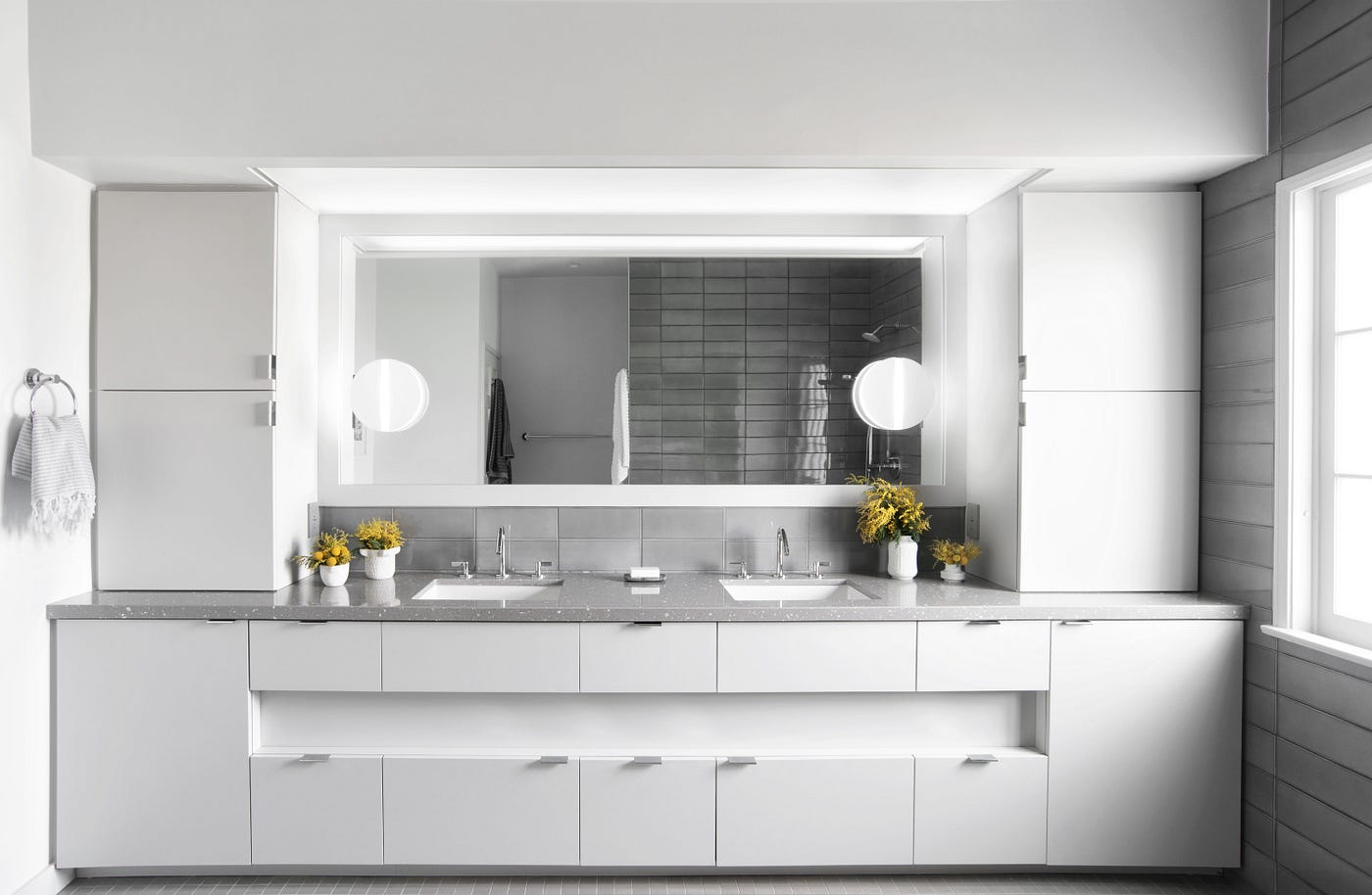 A bright white and grey primary bathroom vanity with clean white custom cabinetry, soft-grey countertops containing pieces of natural shell, grey hand-glazed ceramic backsplash, and a wide mirror with round sconces on each side.
