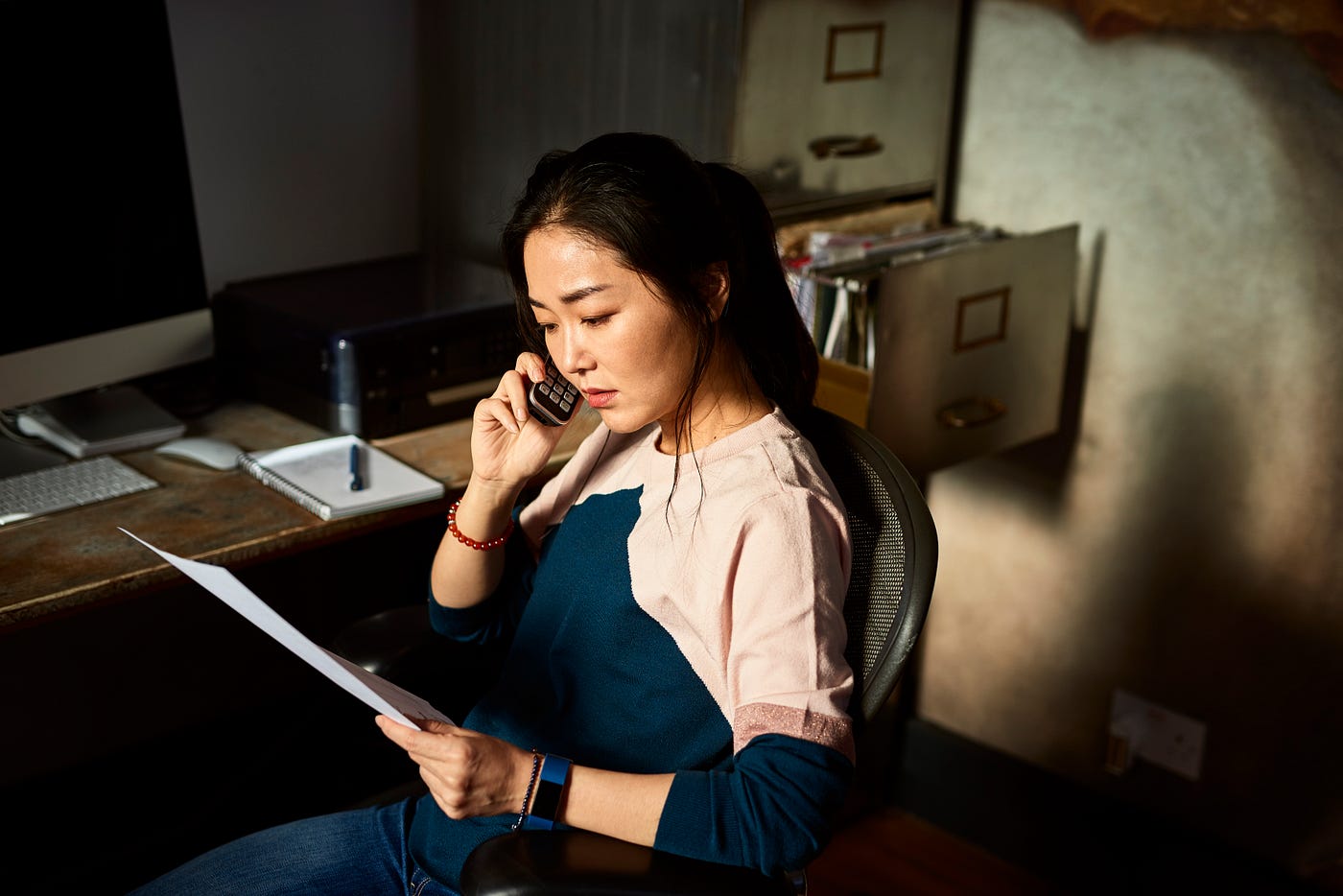 A woman looks serious as she listens on the phone while holding paperwork and sitting in an office chair.