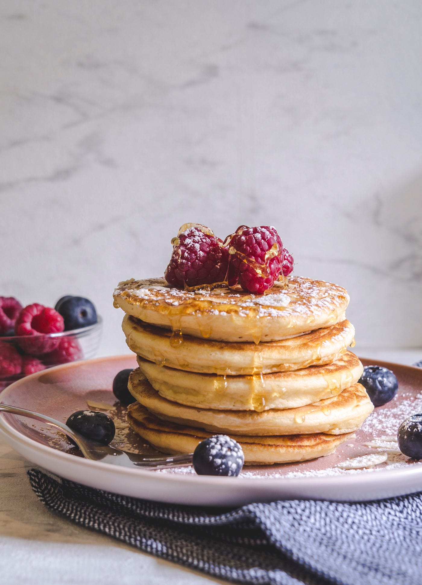 A stack of pancakes with berries and a drizzle of hony on a marble counter top.