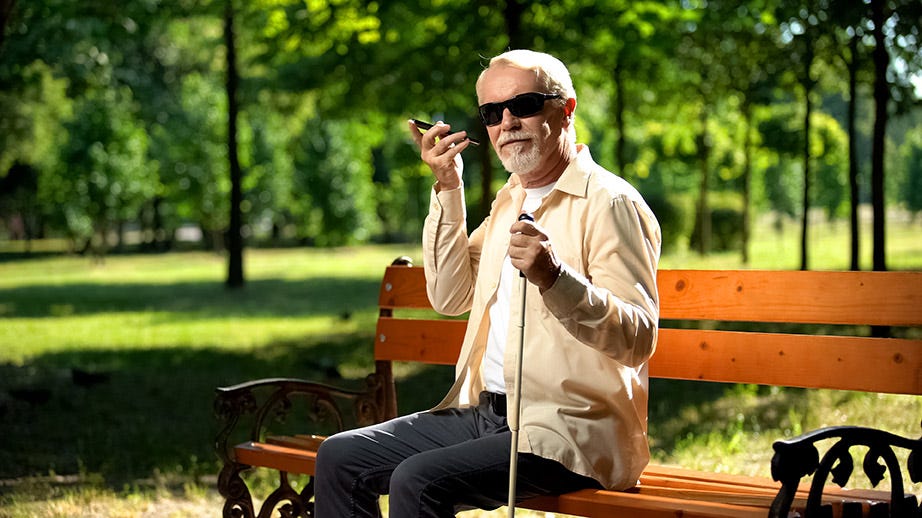 Older man wearing dark glasses and holding a white cane in left hand, sitting on park bench holding smartphone to his ear with right hand.