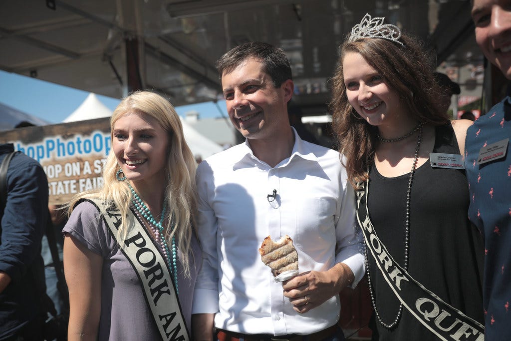 Pete Buttigieg stands holding a pork chop on a stick, accompanied by the Iowa Pork Queen at the Iowa State Fair.