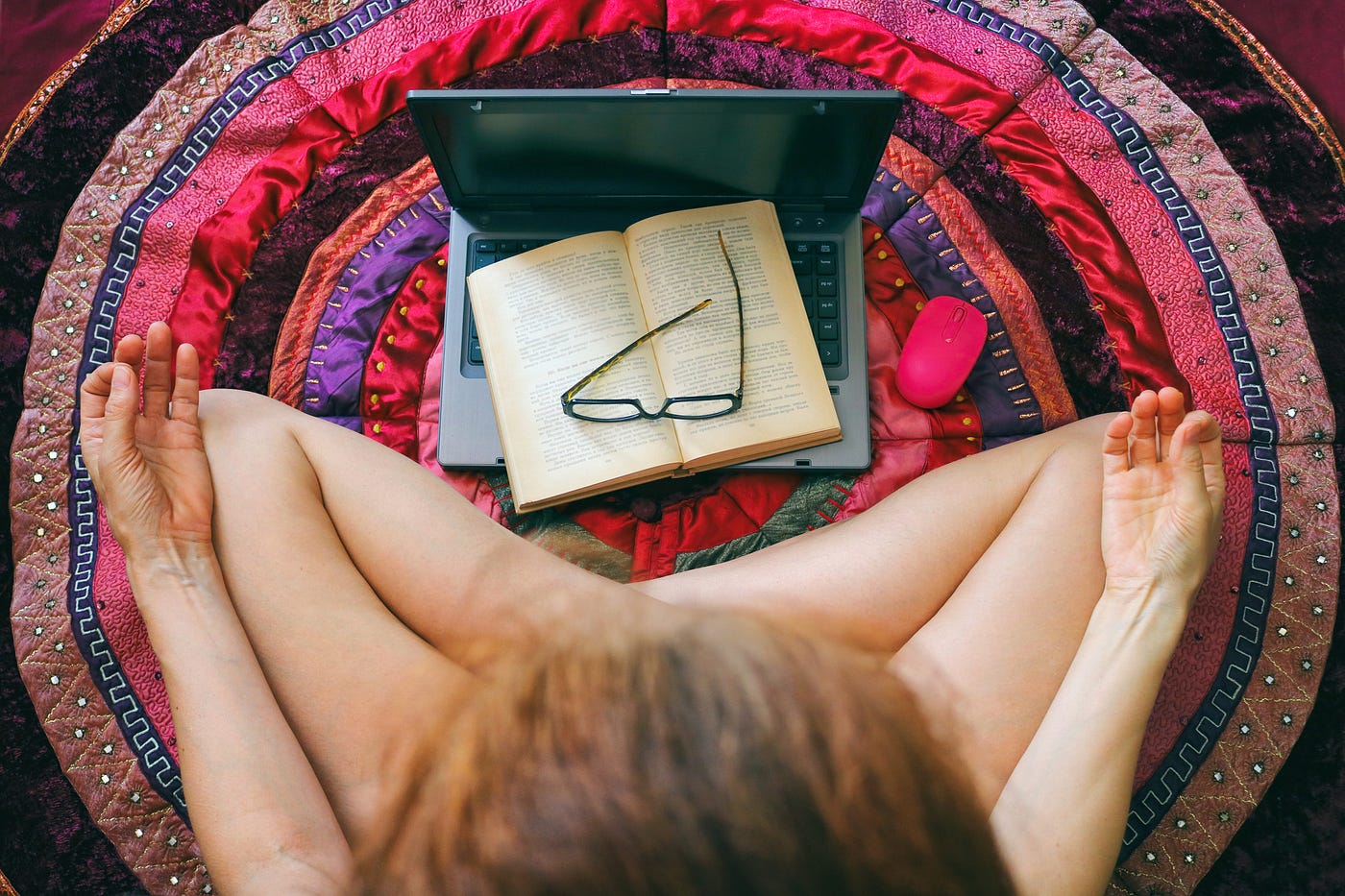 A photo of a woman sitting cross-legged, meditating, on the floor. A laptop with an open book and glasses is in front of her.
