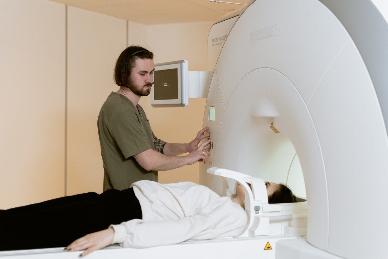 A woman lays on her back in an MRI machine near a male healthcare worker who is operating the machine