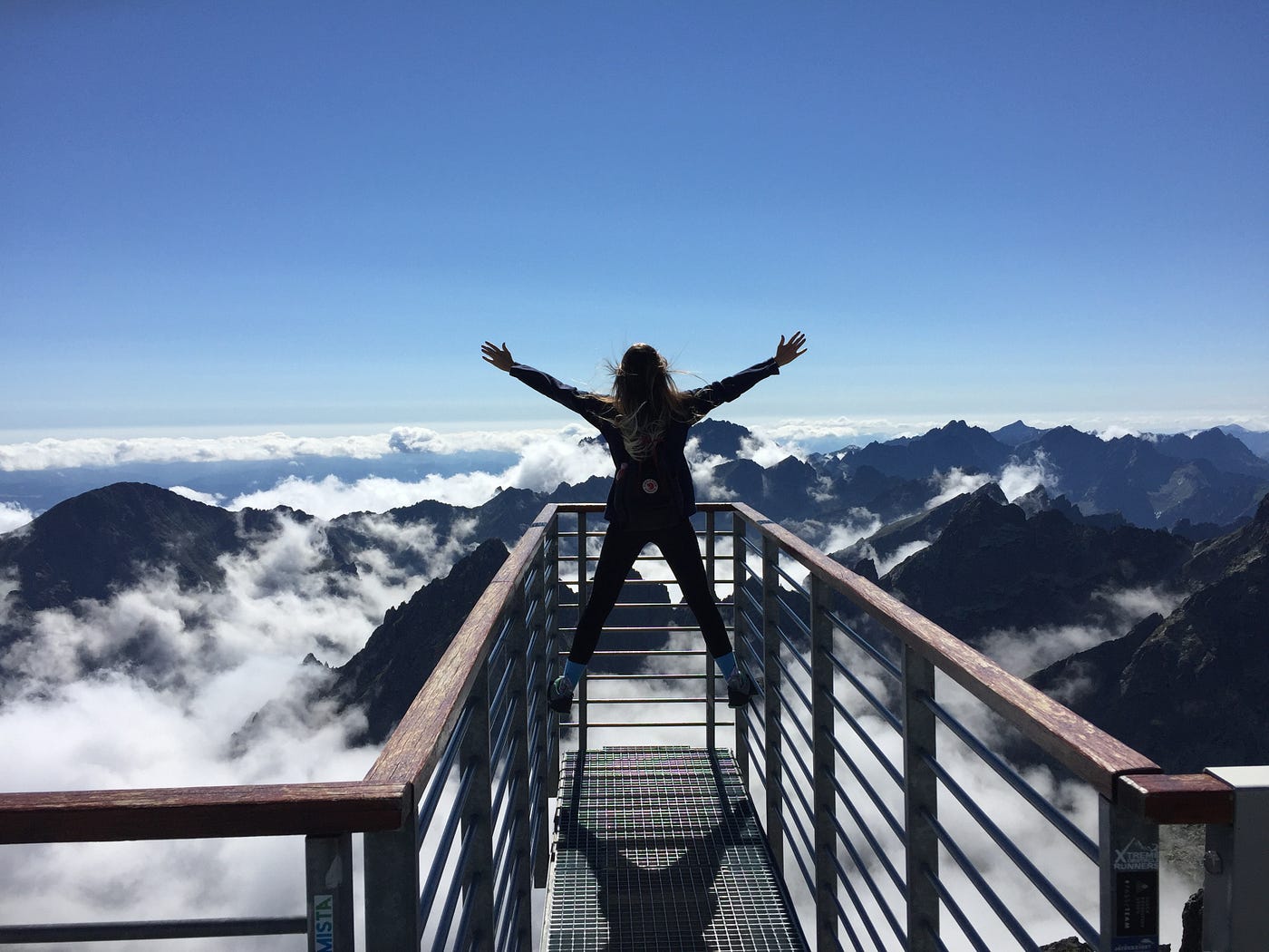 A woman triumphantly stands on a railing viewing the vast cloudy mountains in front of her.