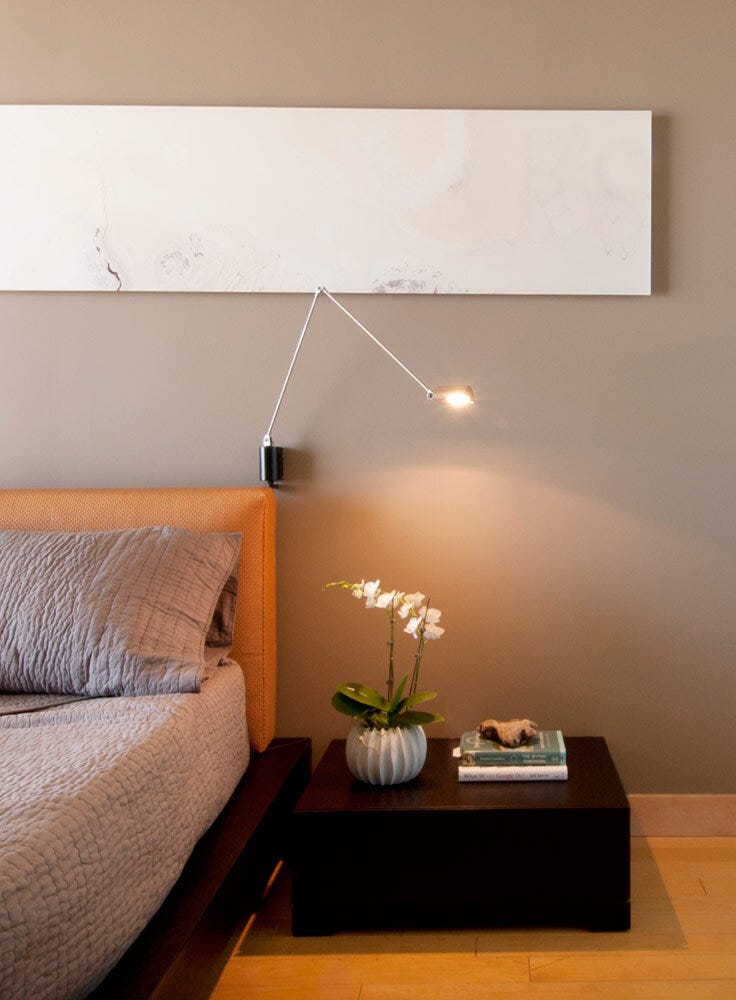 A bed with a brown headboard, a nightstand displaying a book and a plant, and a wall-mounted light.