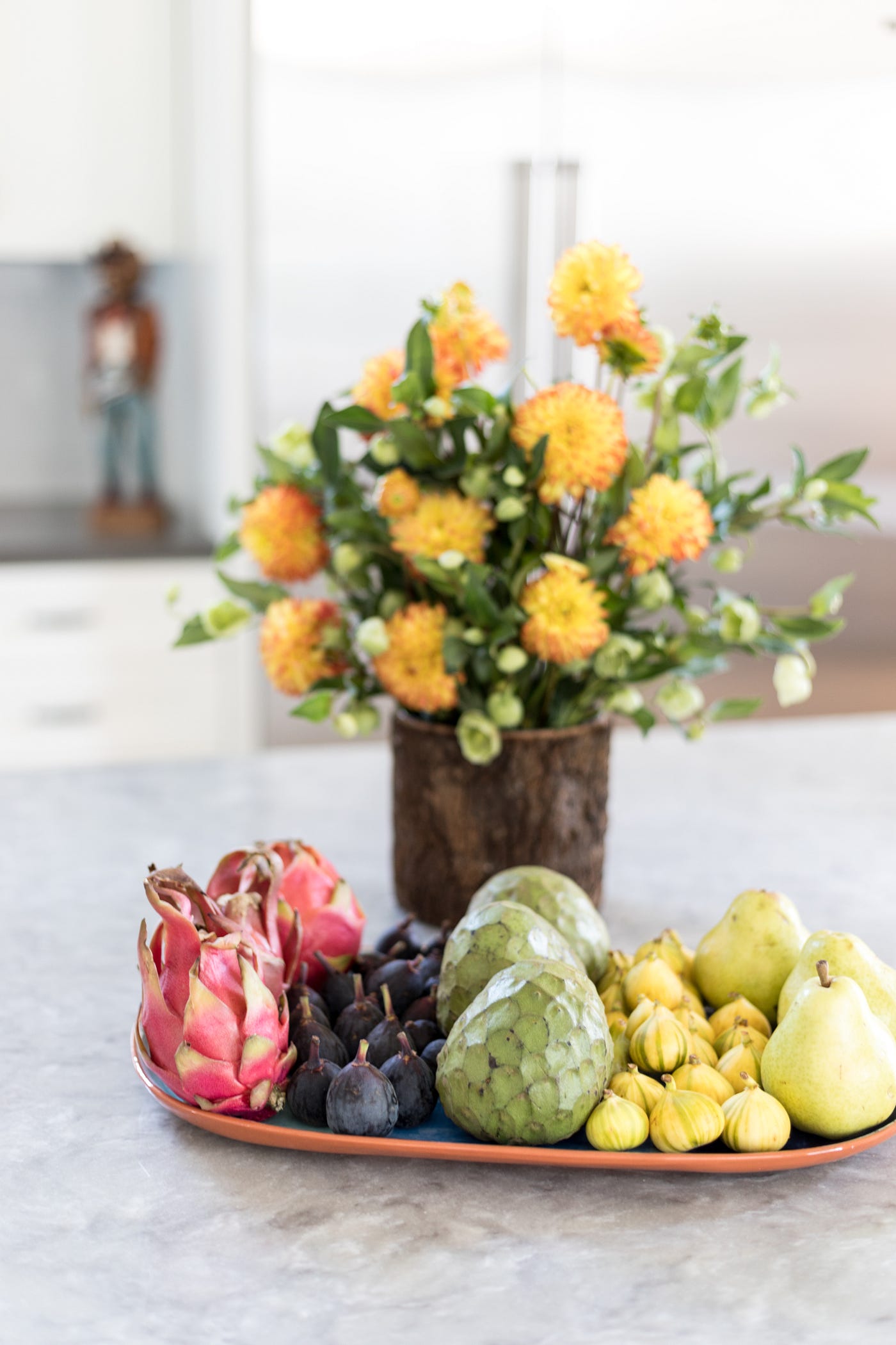 A kitchen countertop with a platter of tropical fruits and a wooden vase with bright yellow flowers.