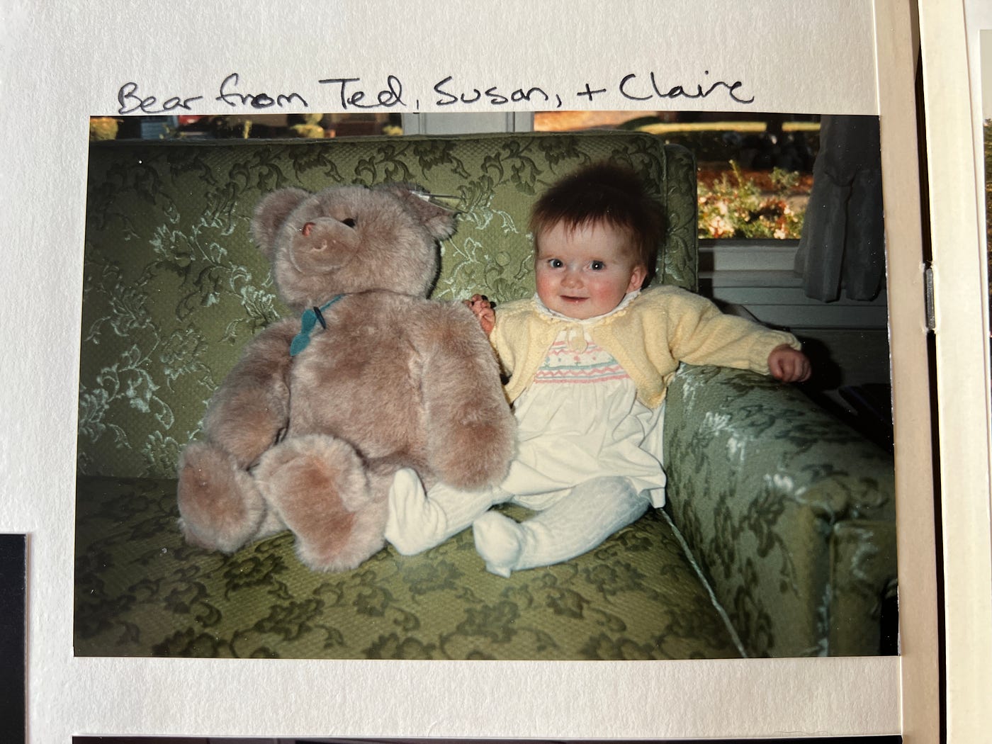 A picture of Victoria in a yellow sweater and stockings next to a giant teddy bear sitting on a couch with flowered green upholstery. Victoria smiles and looks right into camera.