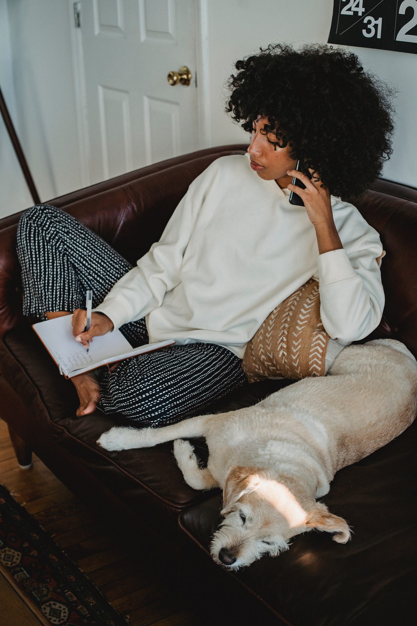Woman sitting on a couch with a dog. She’s holding the phone in one hand and taking notes with the other.