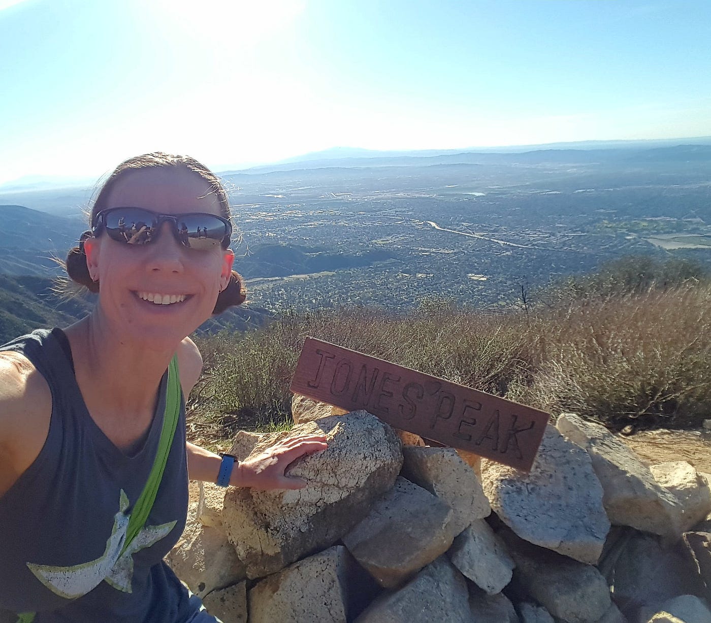 Smiling woman taking selfie with sign on pile of rocks that says, “Jones Peak” and view behind from top of mountain.