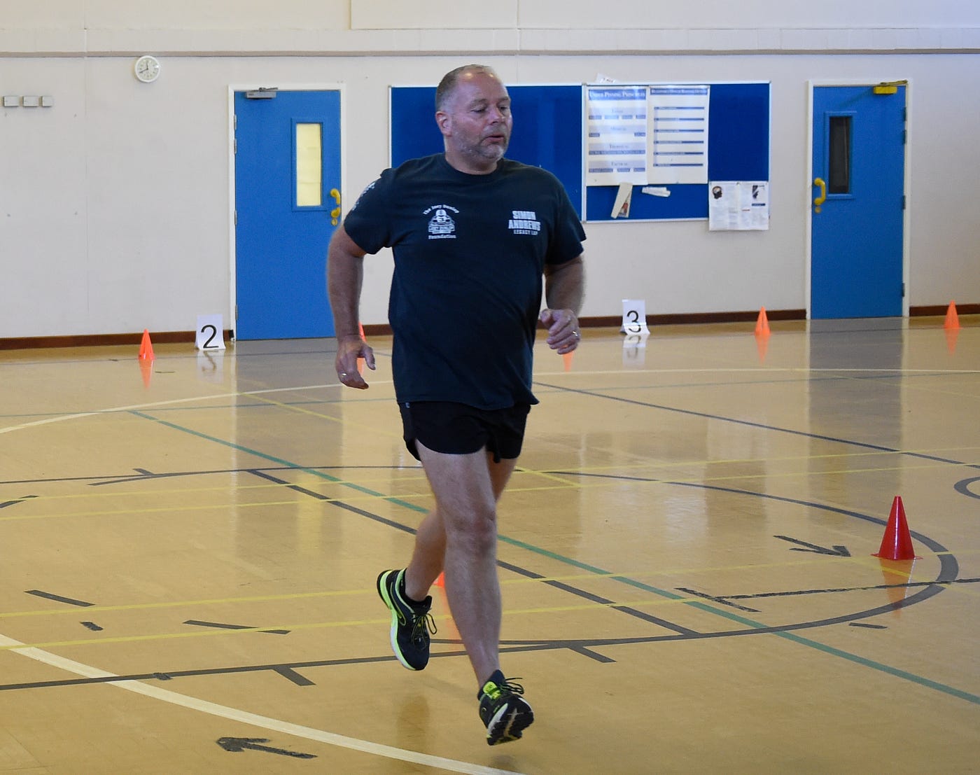 Man wearing sports clothes, running in a sports hall with cones on the floor and blue doors and notice boards in the background