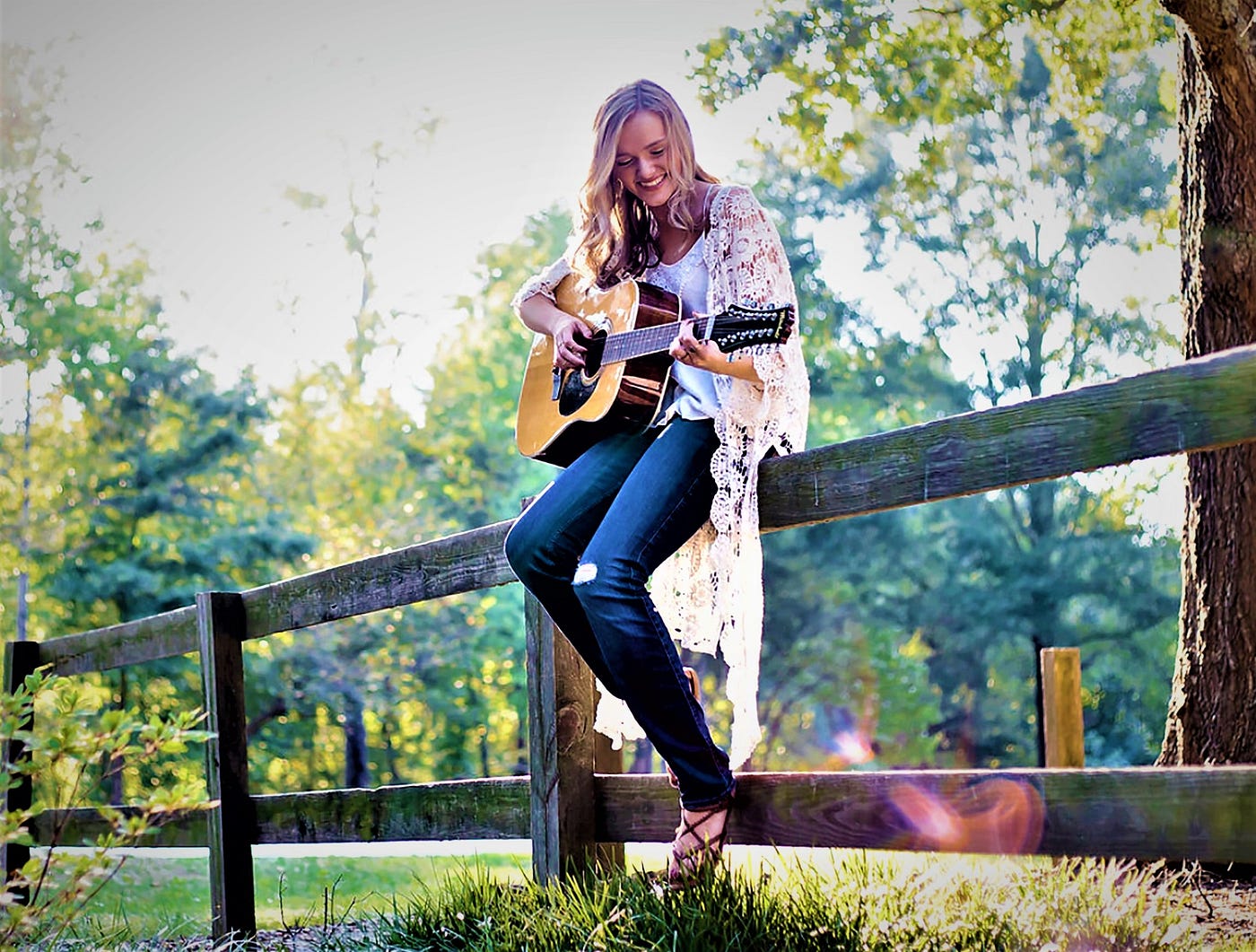 smiling girl with long blonde hair sitting on wooden fence and playing guitar