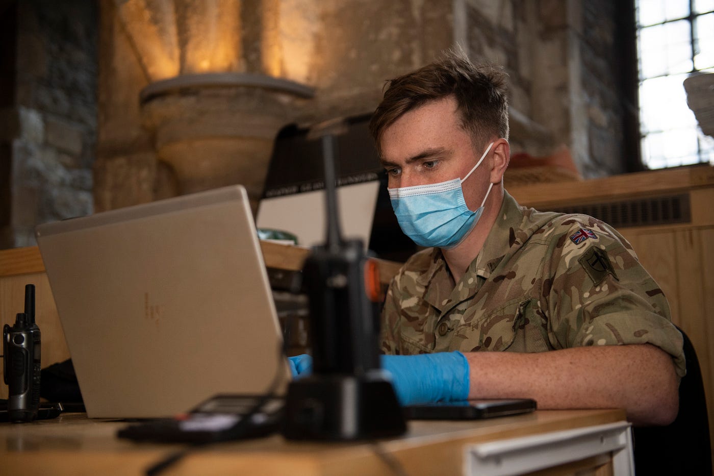 A soldier in a facemask and gloves types into a laptop. There is a walkie-talkie on the desk.