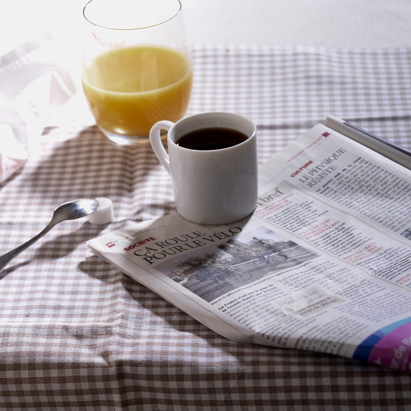 A cup of coffee and a glass of orange juice, on top of a checkered cloth, with a spoon and a newspaper also present. Sylvia.