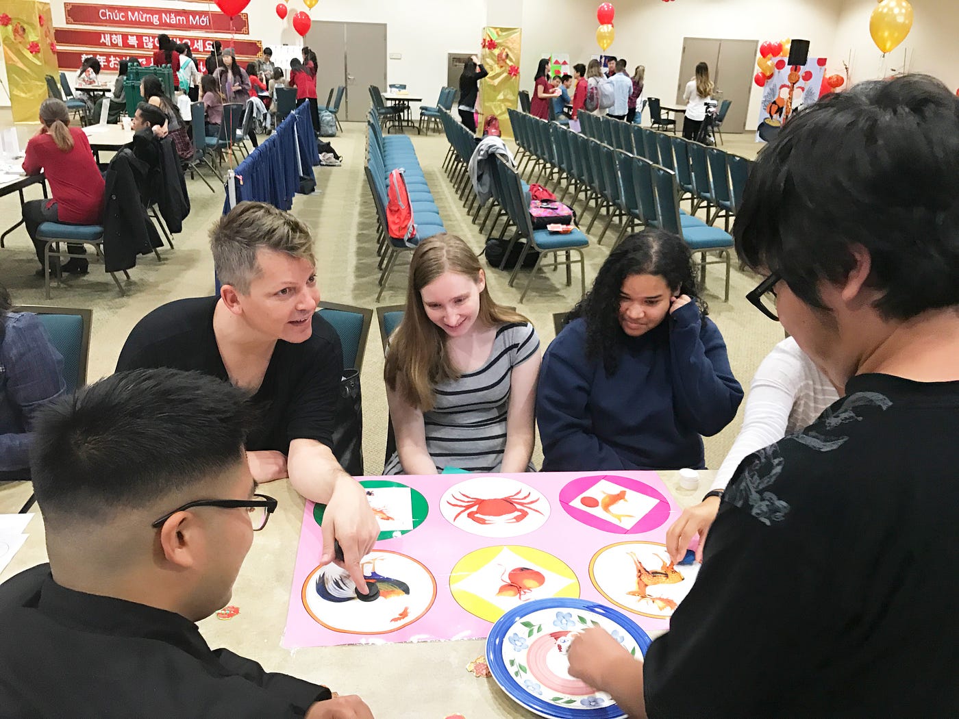 5 people are gathered around a table with circular illustrations playing a game. One man is pointing at a rooster drawing