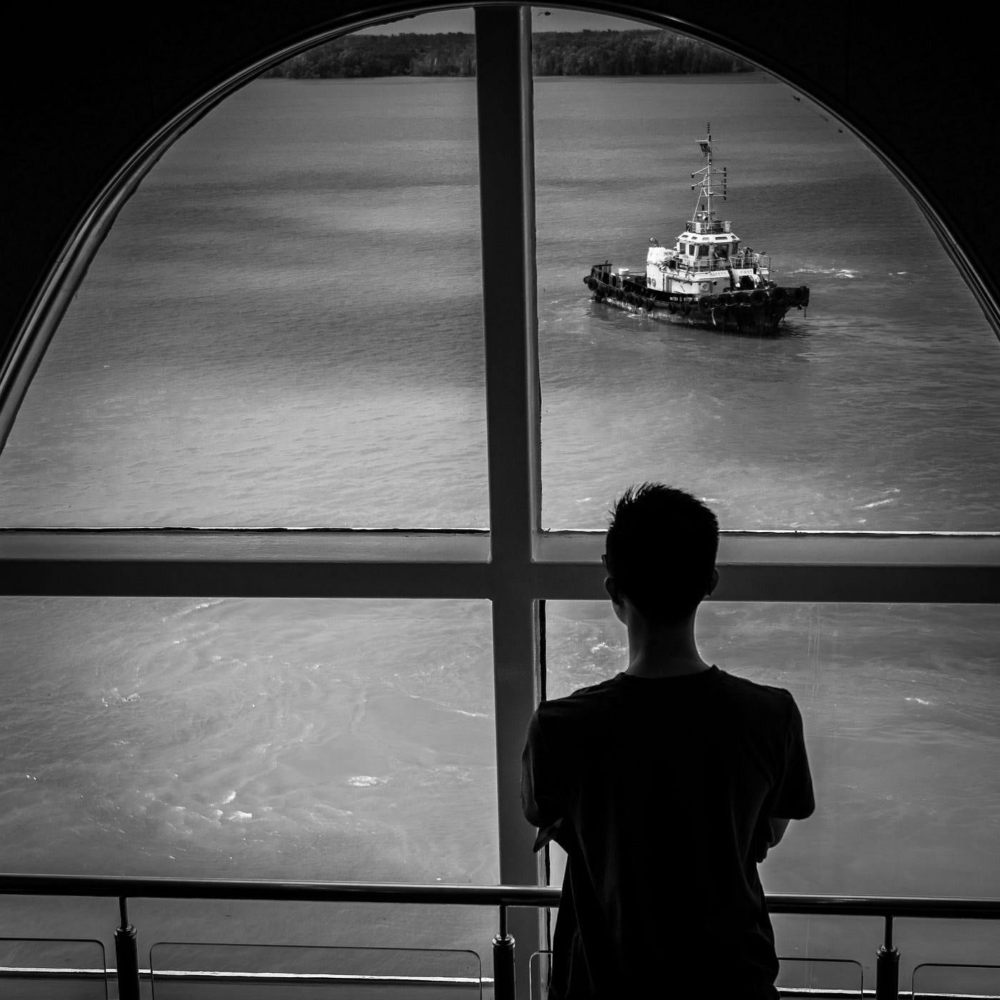 Black and white photo of a person looking through a window. A boat outside.