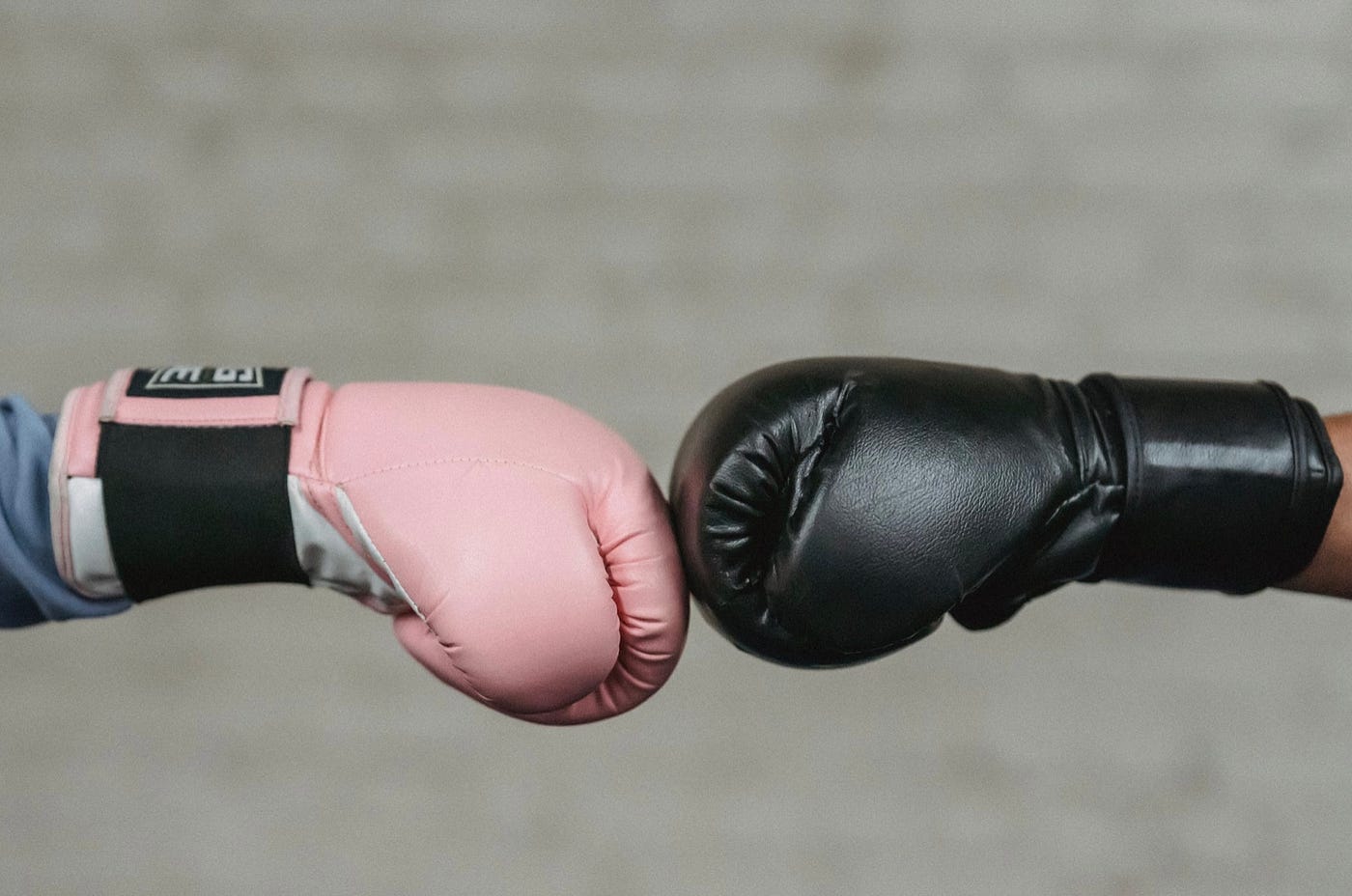 Two boxing gloves are touching at the part of the gloves where the knuckles are held. We see them in profile. The one on the left is pink and the one on the right is black. There is a gray brick wall in the background.
