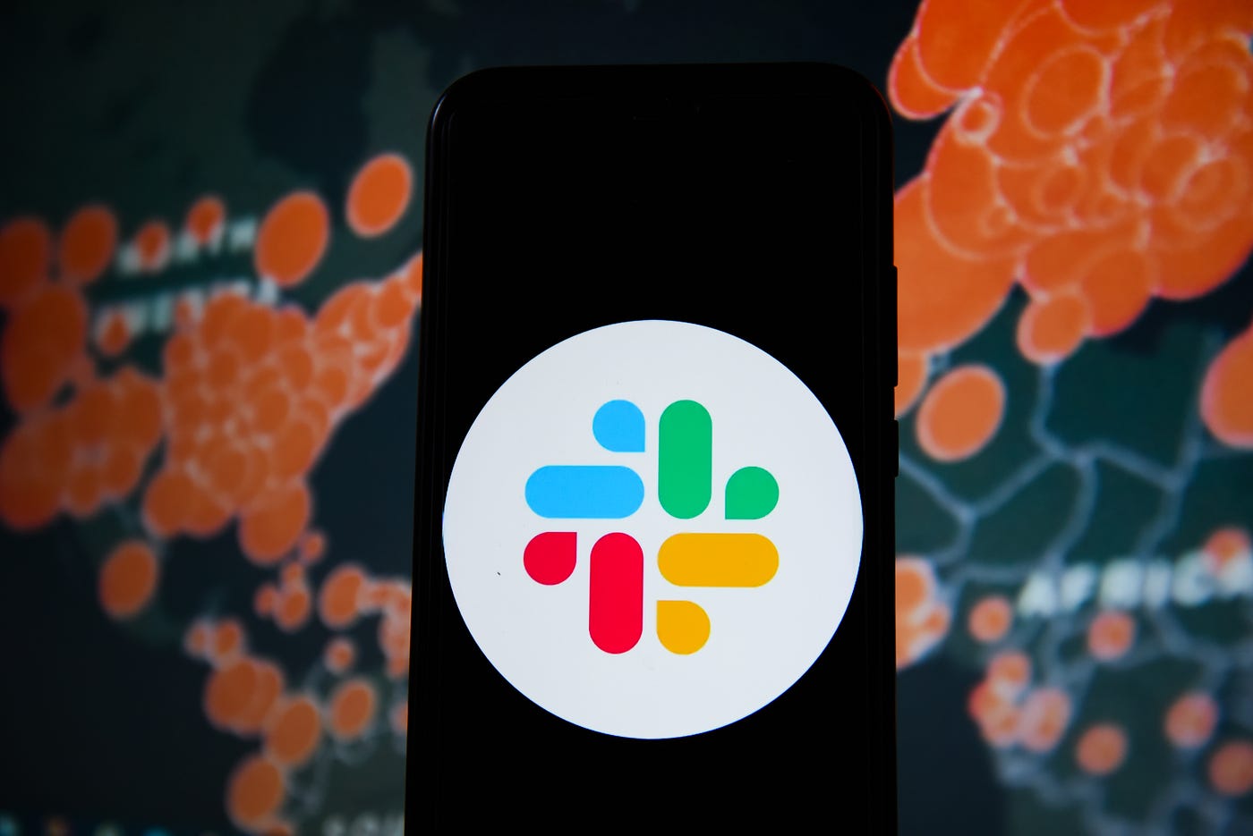 In this photo illustration a Slack logo seen displayed on a phone with a World map of COVID 19 epidemic on the background.