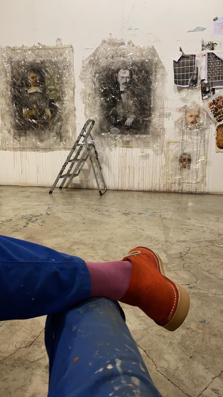 The leg of the photographer is seen in the foreground with his studio and large portraits covered in tape in the background.
