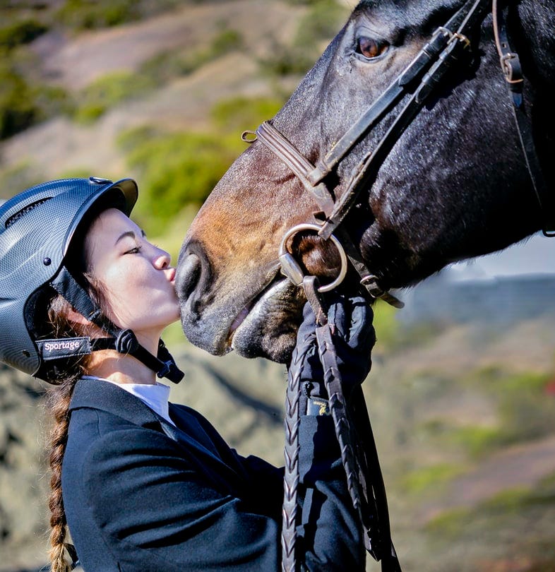 A young woman wearing a helmet showing affection to her horse’s snout