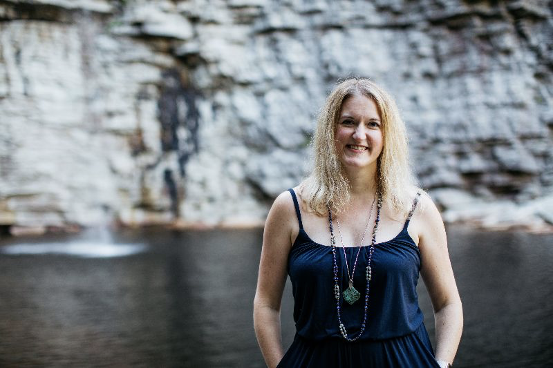 Blonde woman standing in front of a waterfall and lake, rock wall backdrop. Navy blue outfit. Happy, hands in pockets.