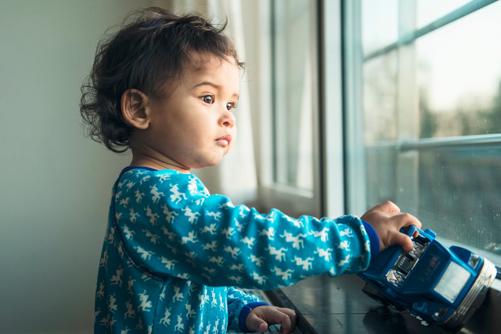 A small child in a aqua jumper holding a blue truck and staring out the window