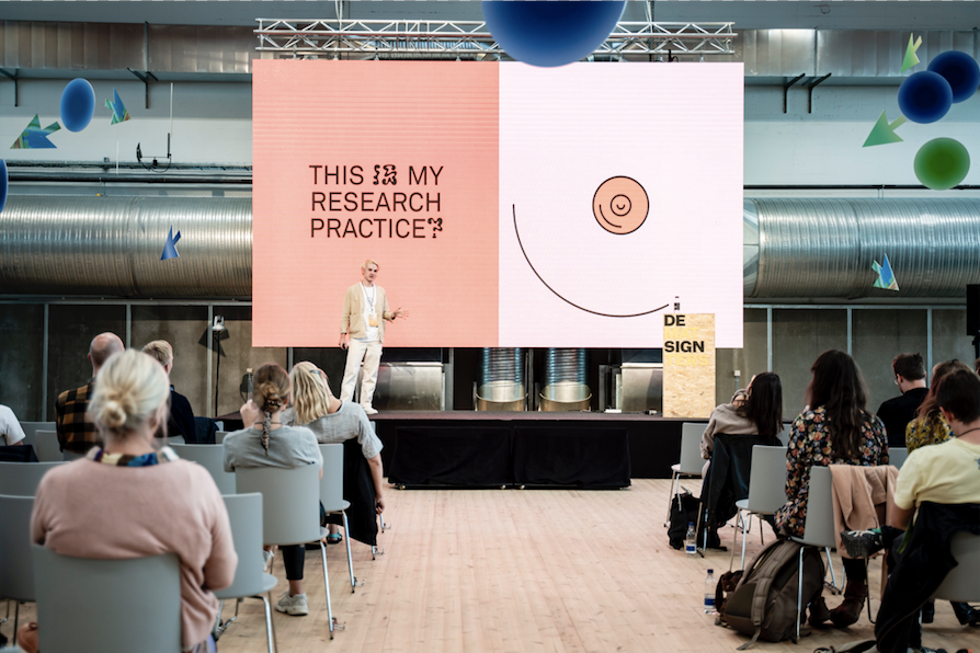 A man on a stage in front of an audience. Behind him a screen says ‘this is my research practice?’ with an abstracted nipple