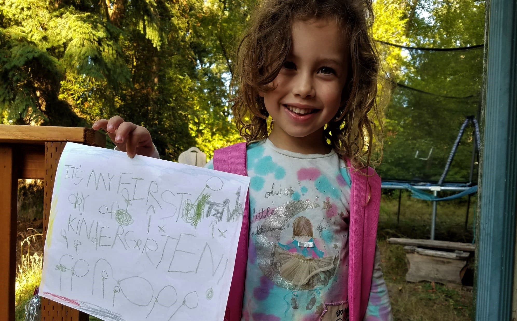 Smiling 5-year-old in a tie-dye shirt and backpack holds a hand-drawn sign that says, “It’s my first day of kindergarten!”