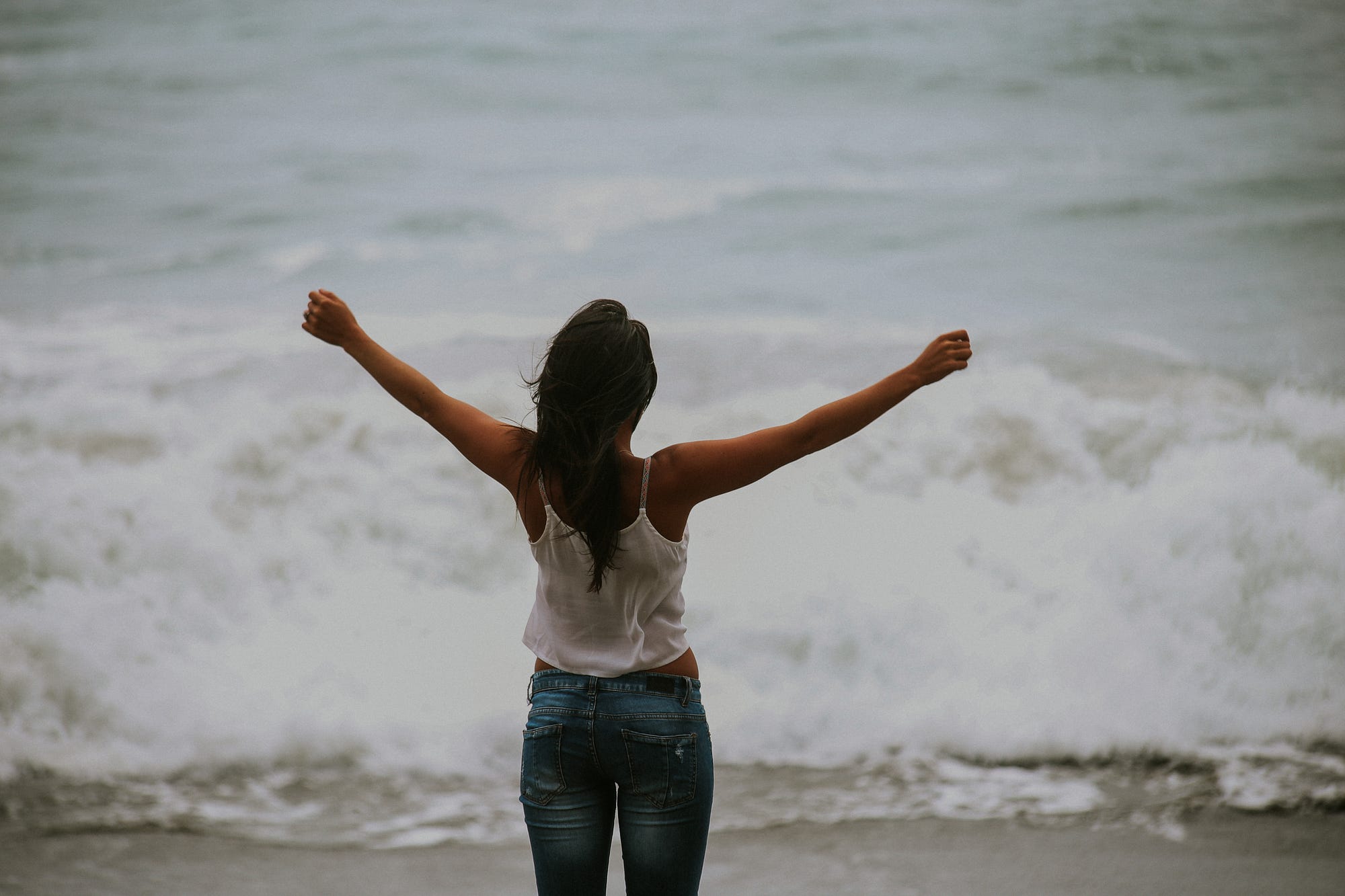 Woman standing on beach in front of waves. Arms open.