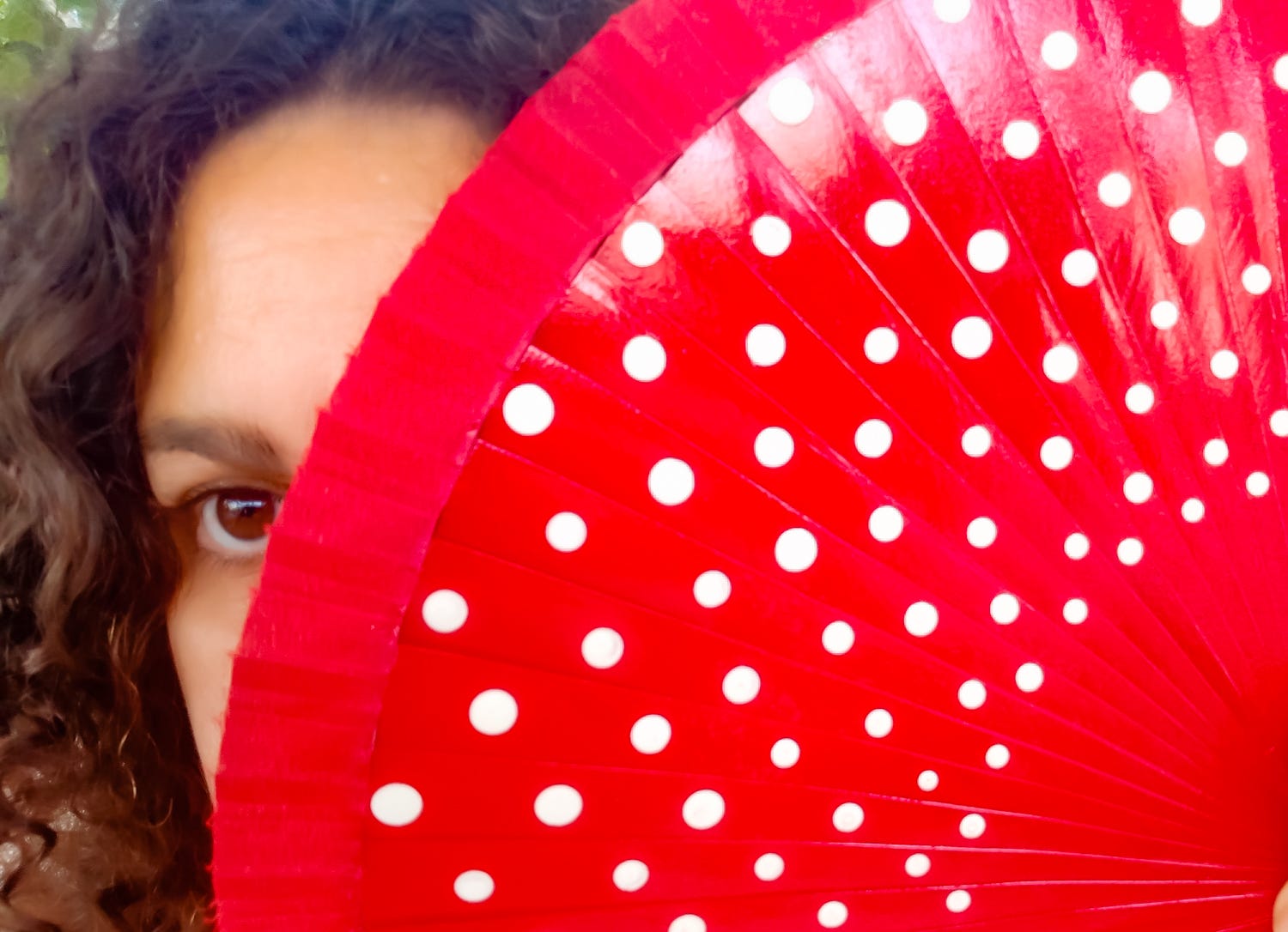 Image of the author partially covering her face to reveal only the left eye looking straight, while hiding behind a Spanish red fan. Her (my) features are Mediterranean, brown eyes, tan skin, and brown curly hair.