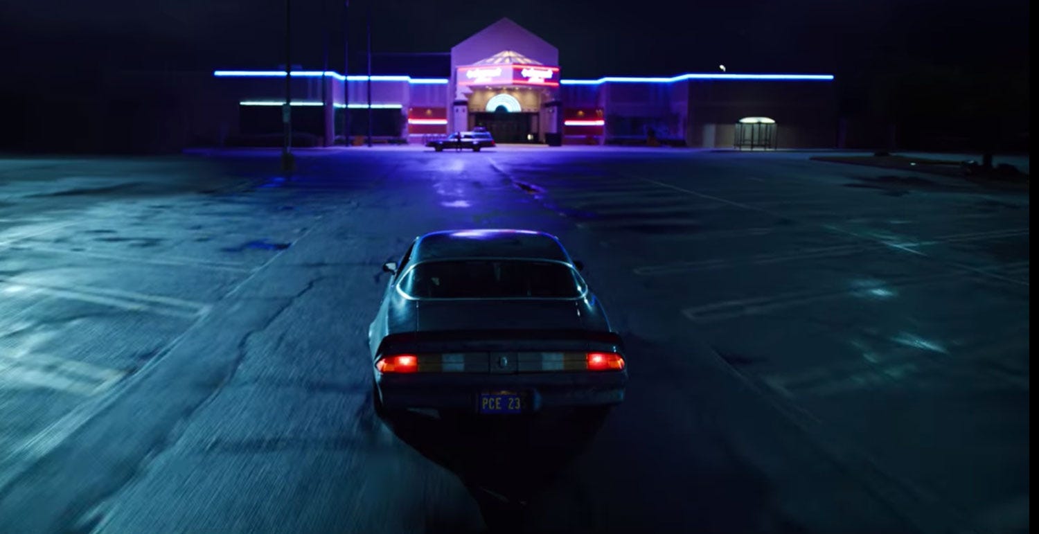 A still from the Stranger Things series on Netflix. A Camaro faces the Starcourt Mall in an empty parking lot at night.