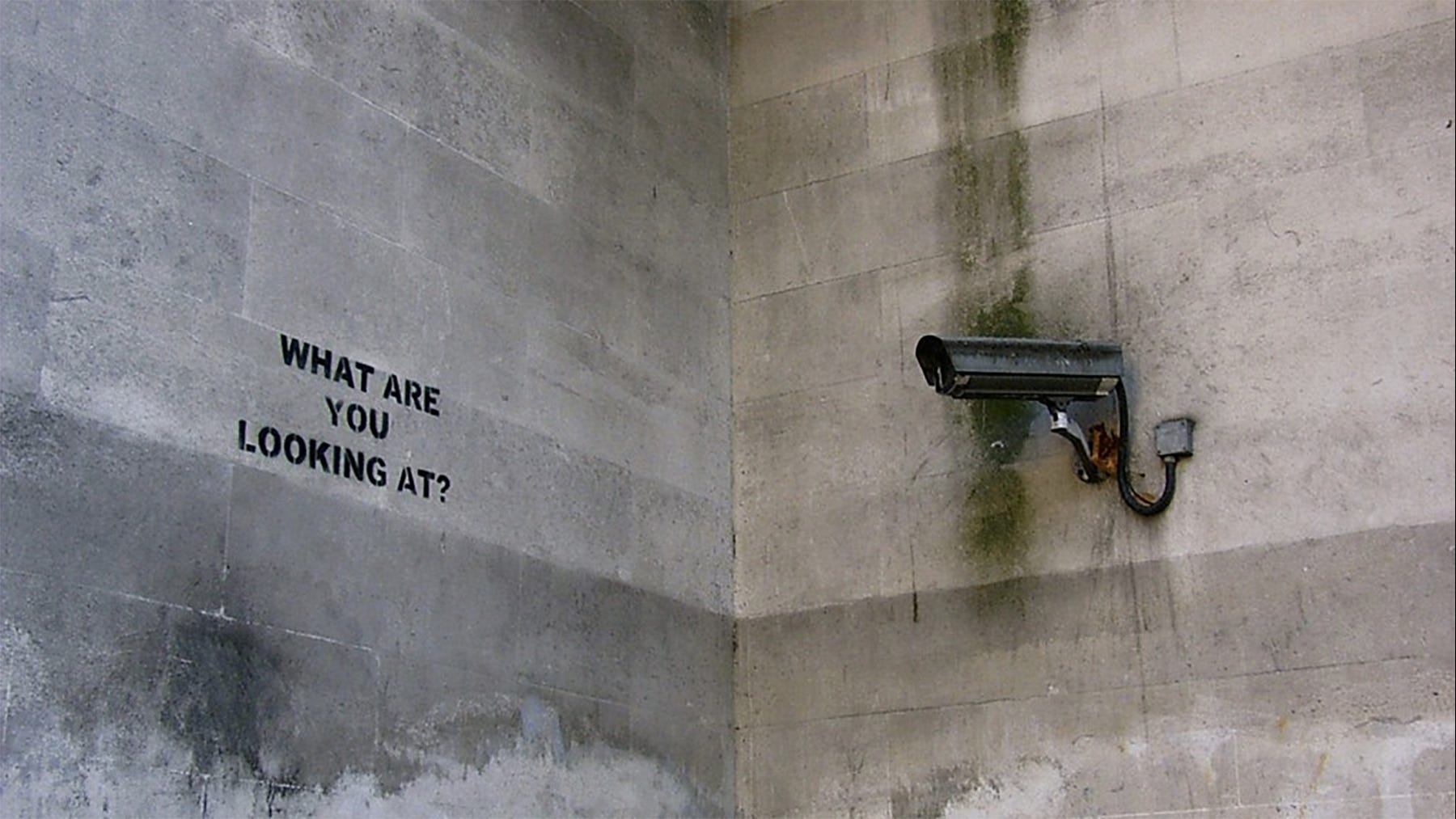Photo of Banksy’s street art reading “What are you looking at?” beside a CCTV camera on a concrete wall.