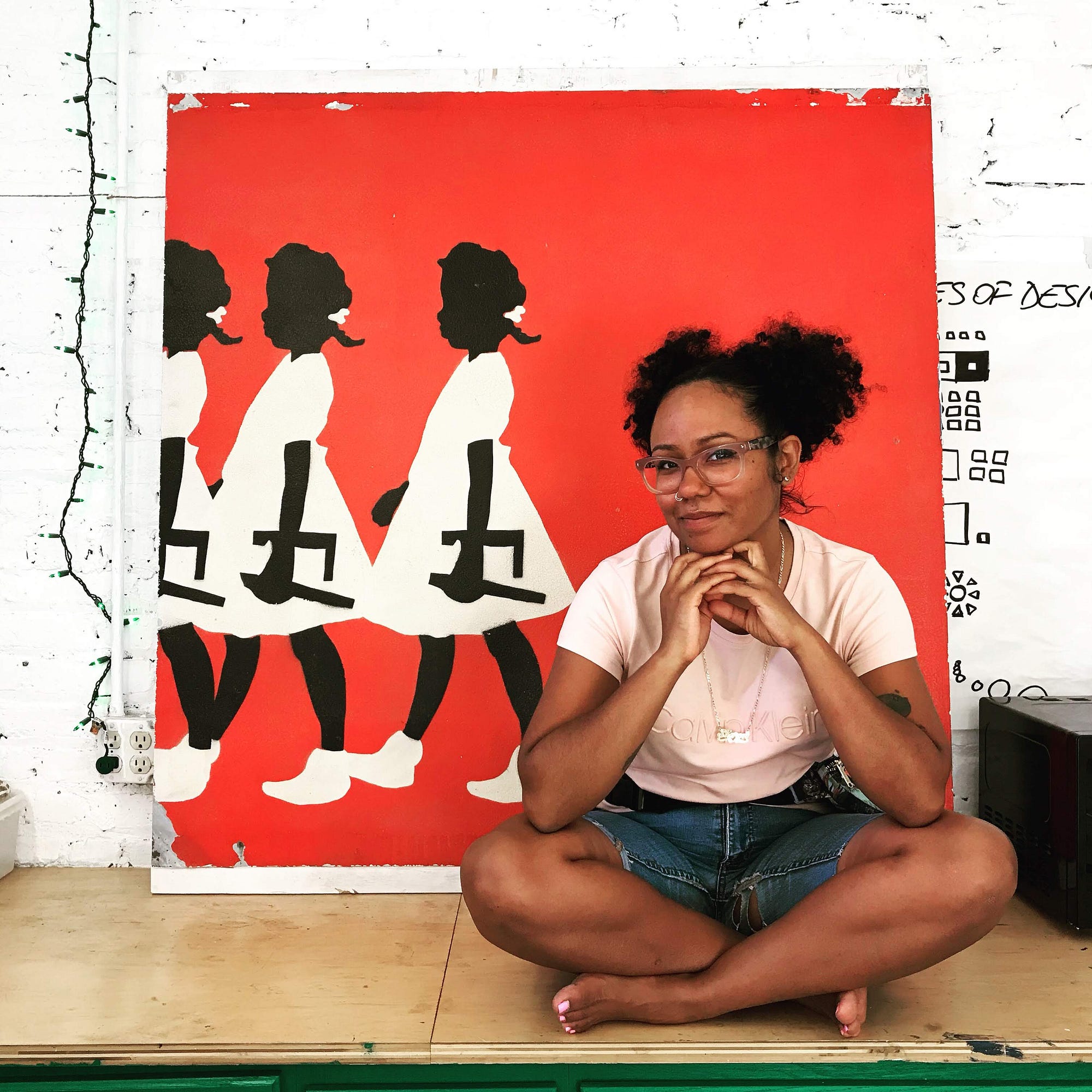 A smiling woman sits on the floor in front of a bright painting featuring three Black girls. Sharon has black hair & glasses