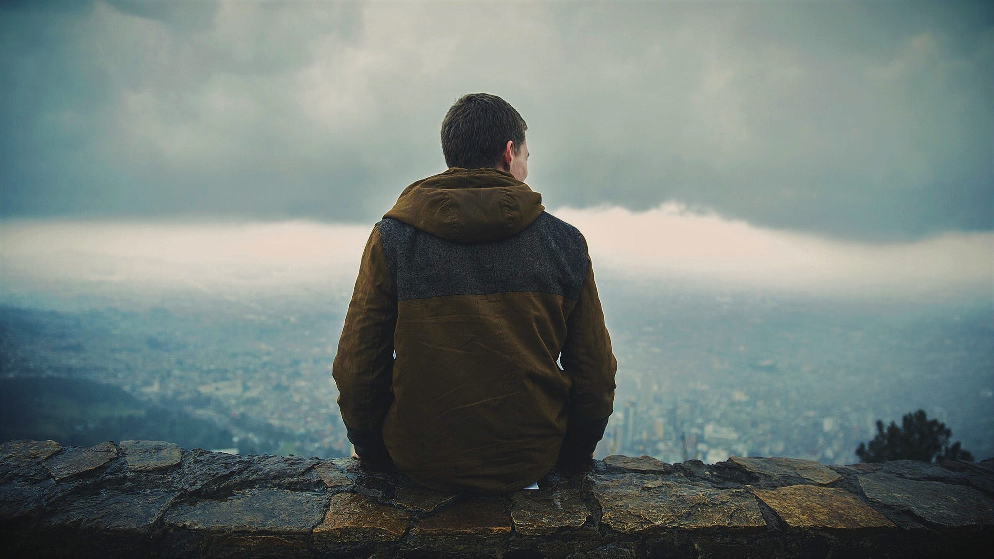 man in hooded jacket sitting on stone wall looking out over landscape and stormy sky
