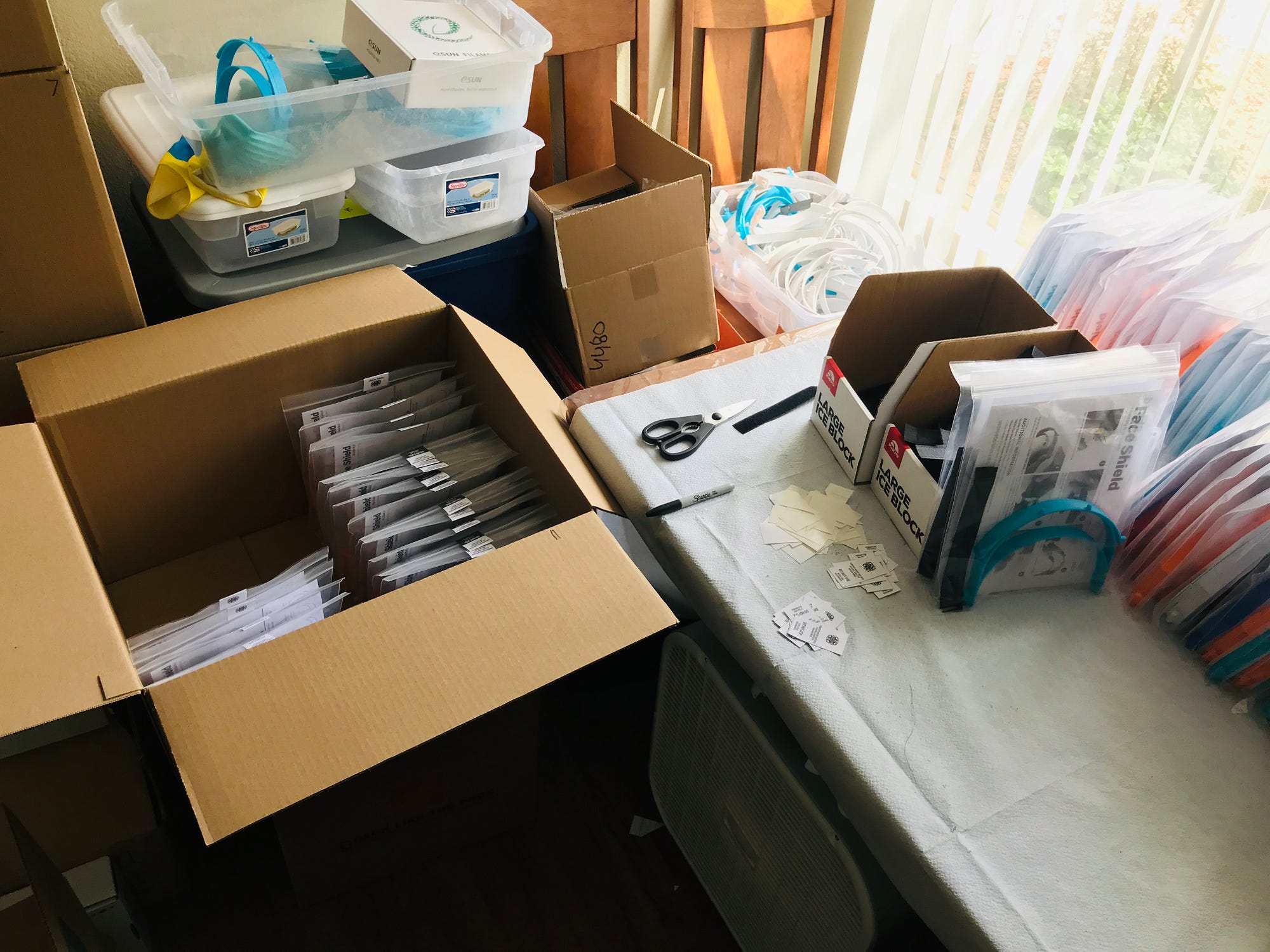 A workspace area with dozens of 3D-printed orange & blue headbands packaged & a box half-full of packaged face shields.
