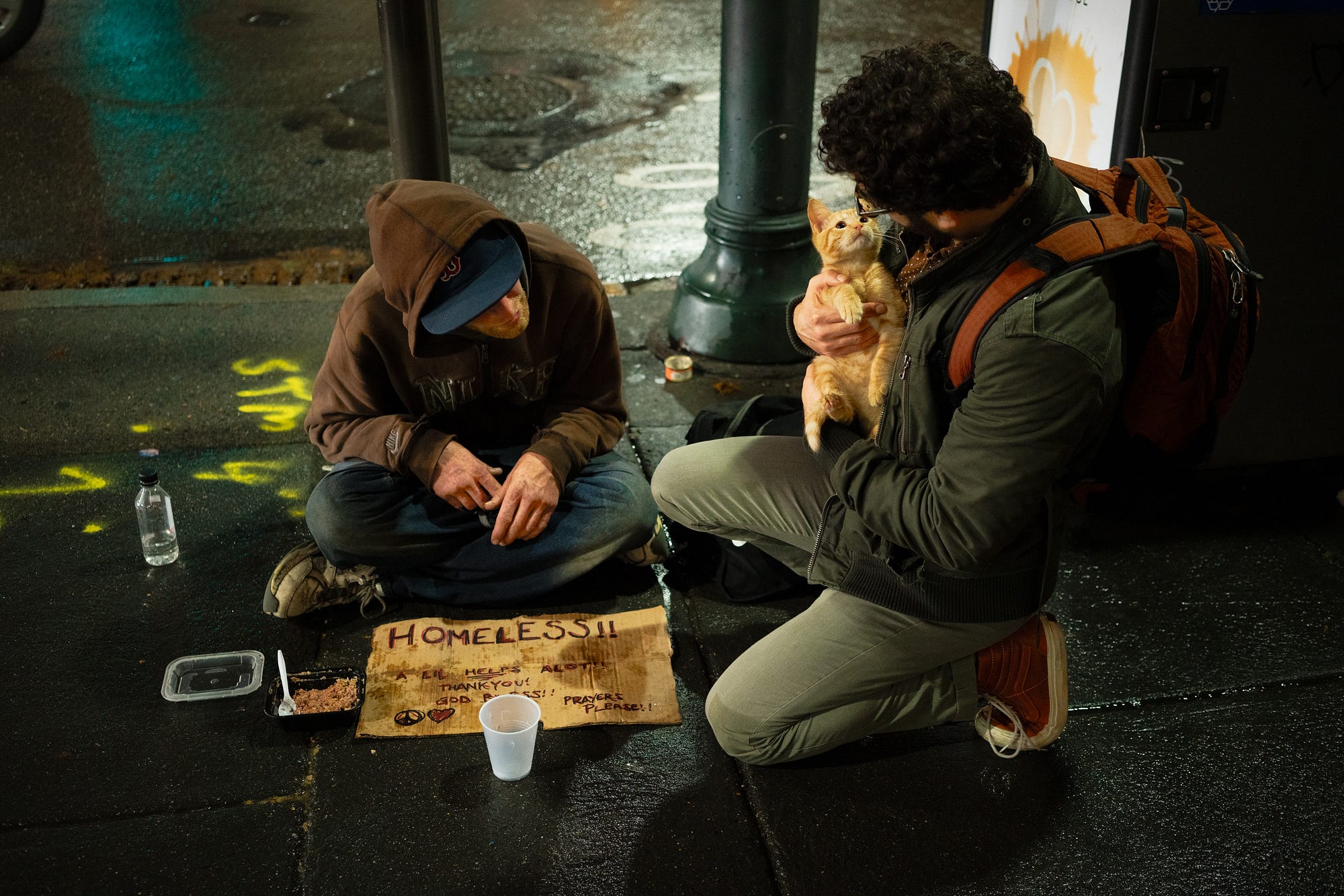 A man wearing a backpack holds an orange cat as he kneels next to a homeless person