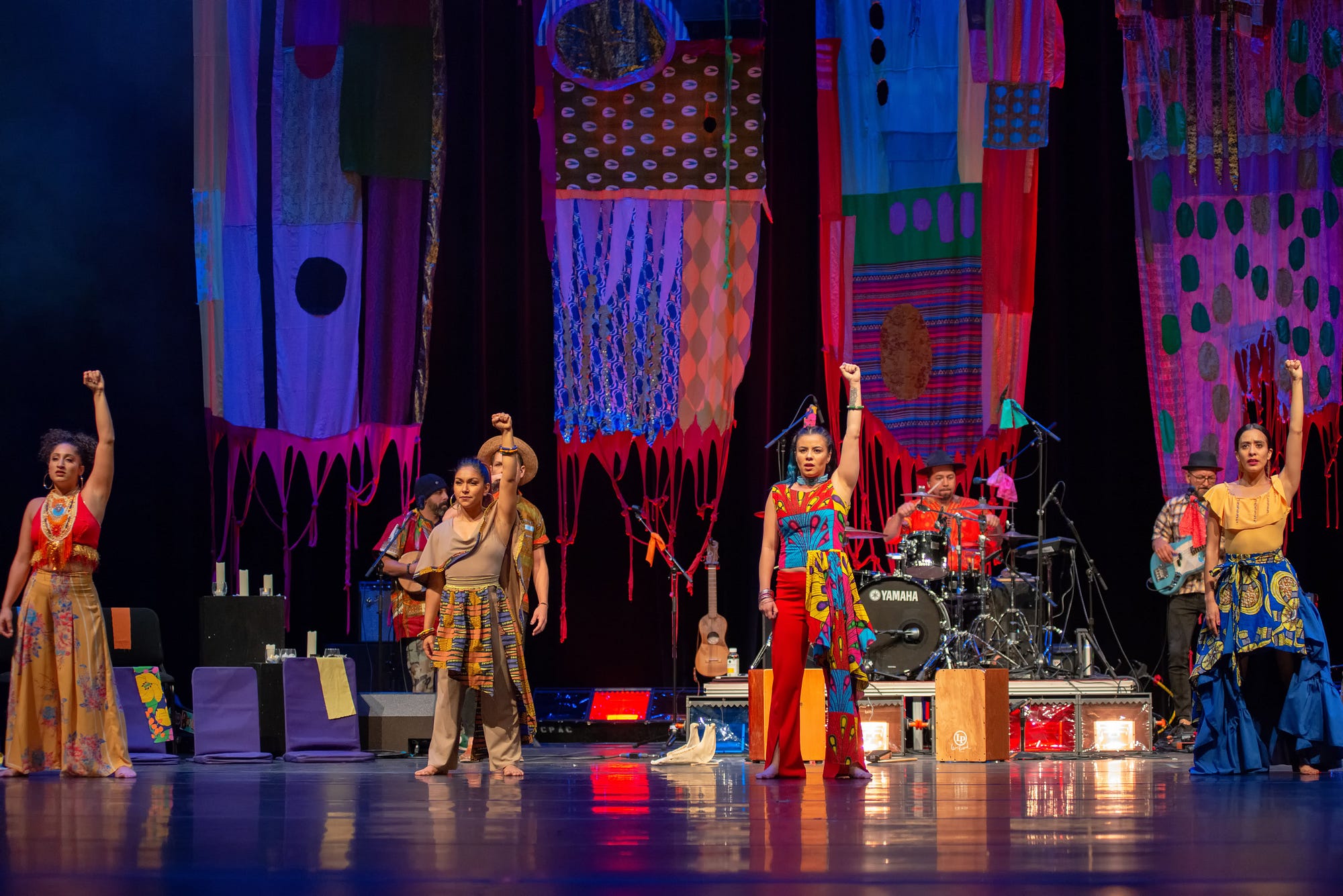 Four women in multicolored dresses stoop forward on a dark stage