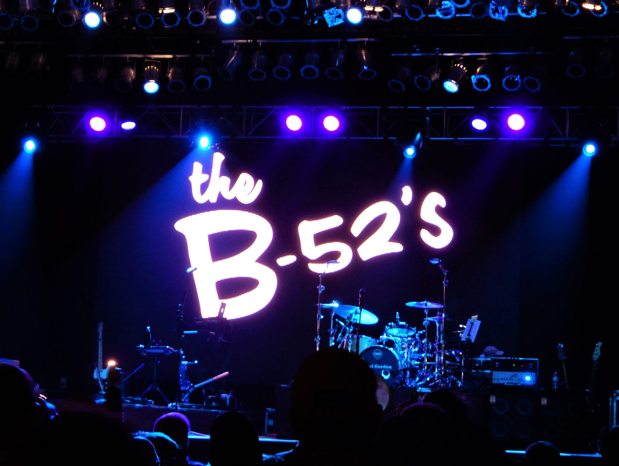 Stage with lit with spotlights and projection of B-52’s logo behind drum kit.