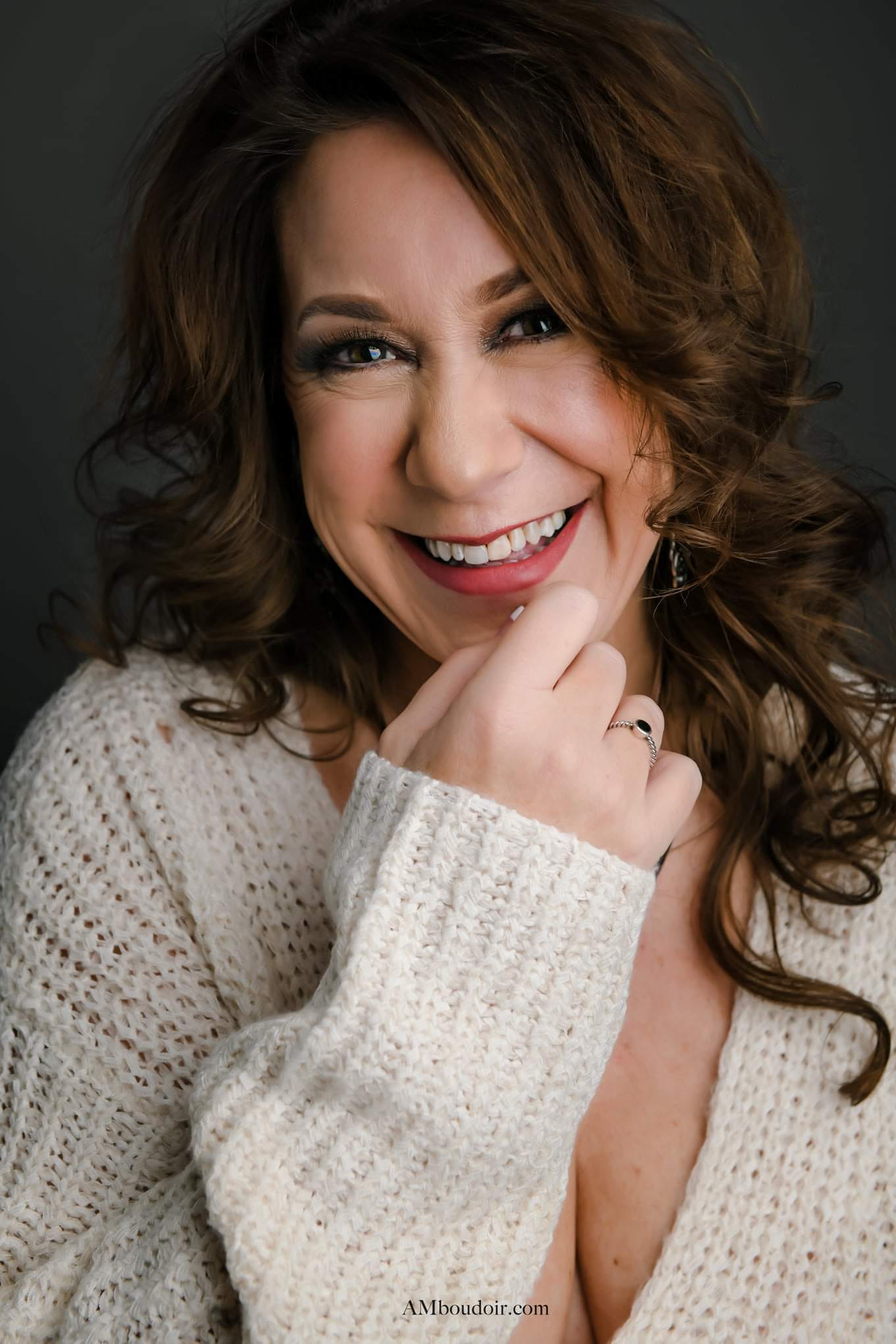 White woman smiling with beige sweater hand at her mouth brown hair curly mauve lipstick and dark eye shadow with brown eye