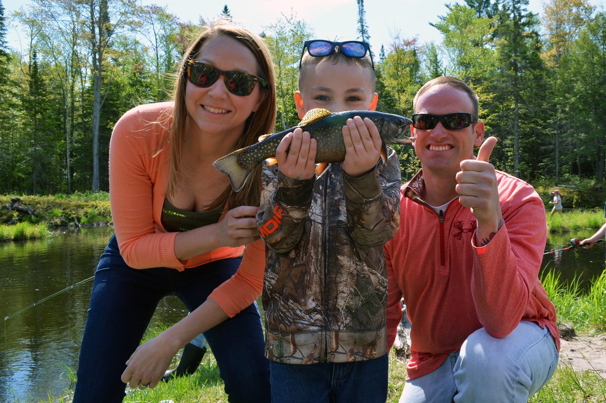 A woman and man pose with a child who is holding a fish. A pond and woods are in the background.