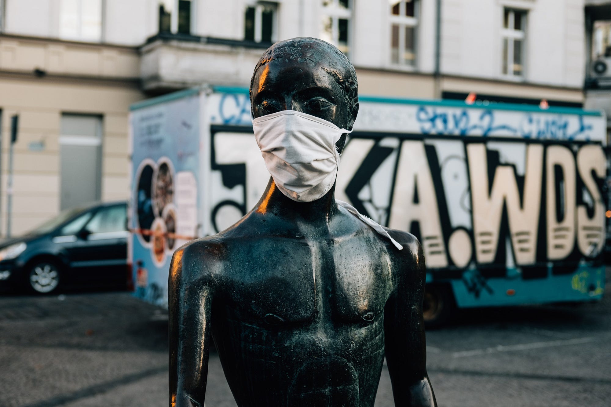 This street scene is of a statue made out of obsidian or something that is ebony, and it is wearing a mask. In the background there is a container like a trailer covered with graffiti.