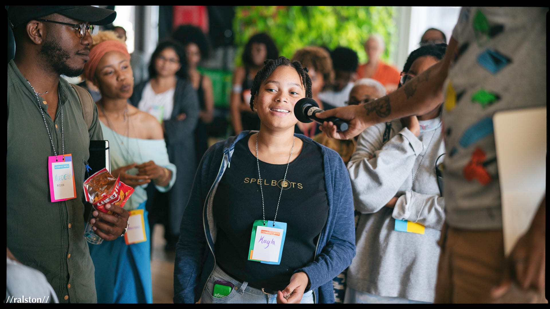 A young, smiling Black person is in a group being interviewed. The surrounding people wear badges that say Afrotectopia.