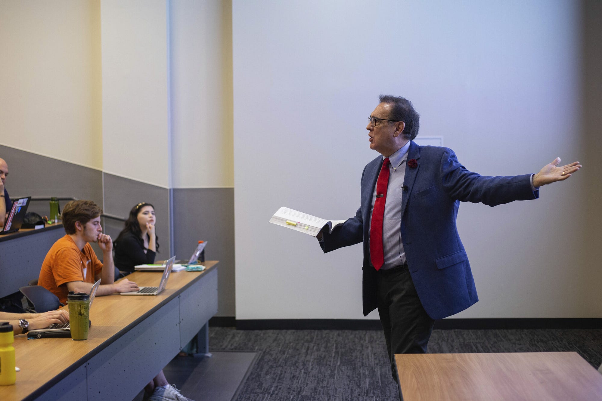 Thomas Garza stands in the front of a classroom of students holding a book and gesturing to the screen behind him.