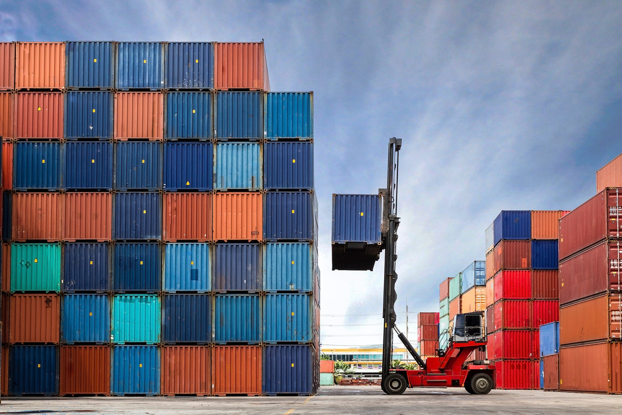 A crane lifting a shipping container next to a tall stack of other containers in a shipping yard.