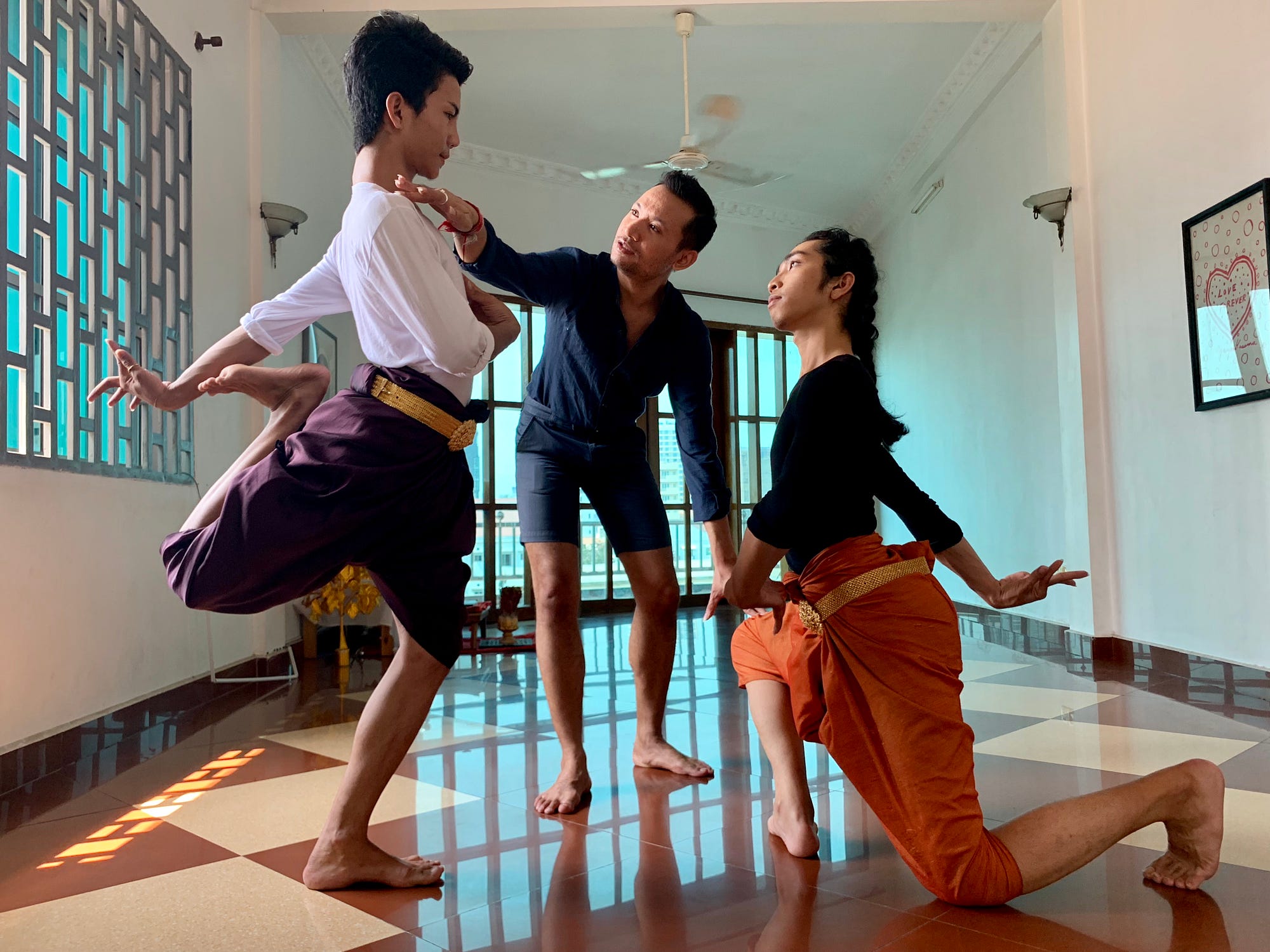 Man instructs two male dancers in Cambodian dance in a dance studio