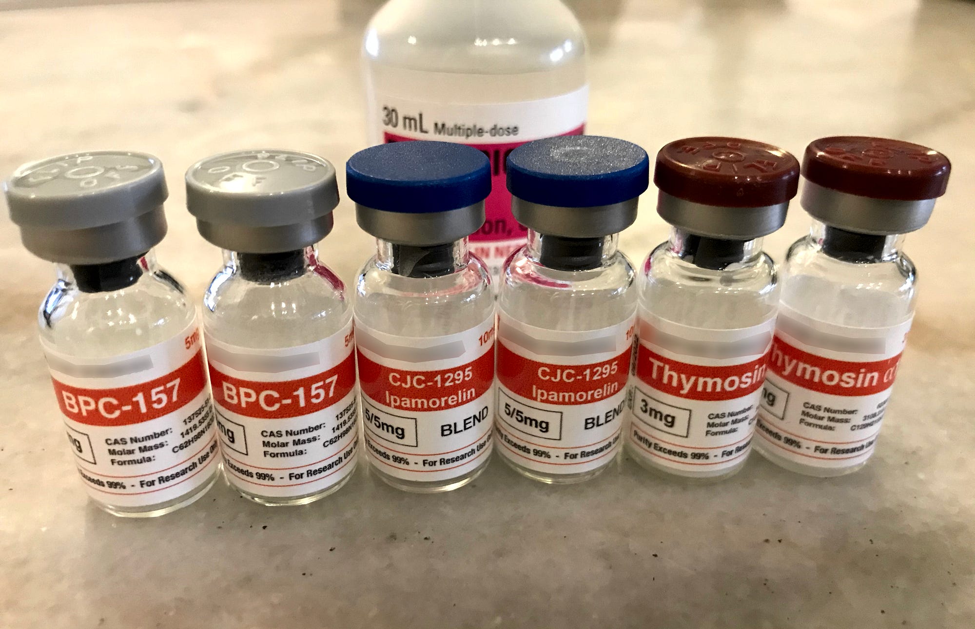 6 small, unopened peptide vials on a table with a bottle of bacteriostatic water behind them.