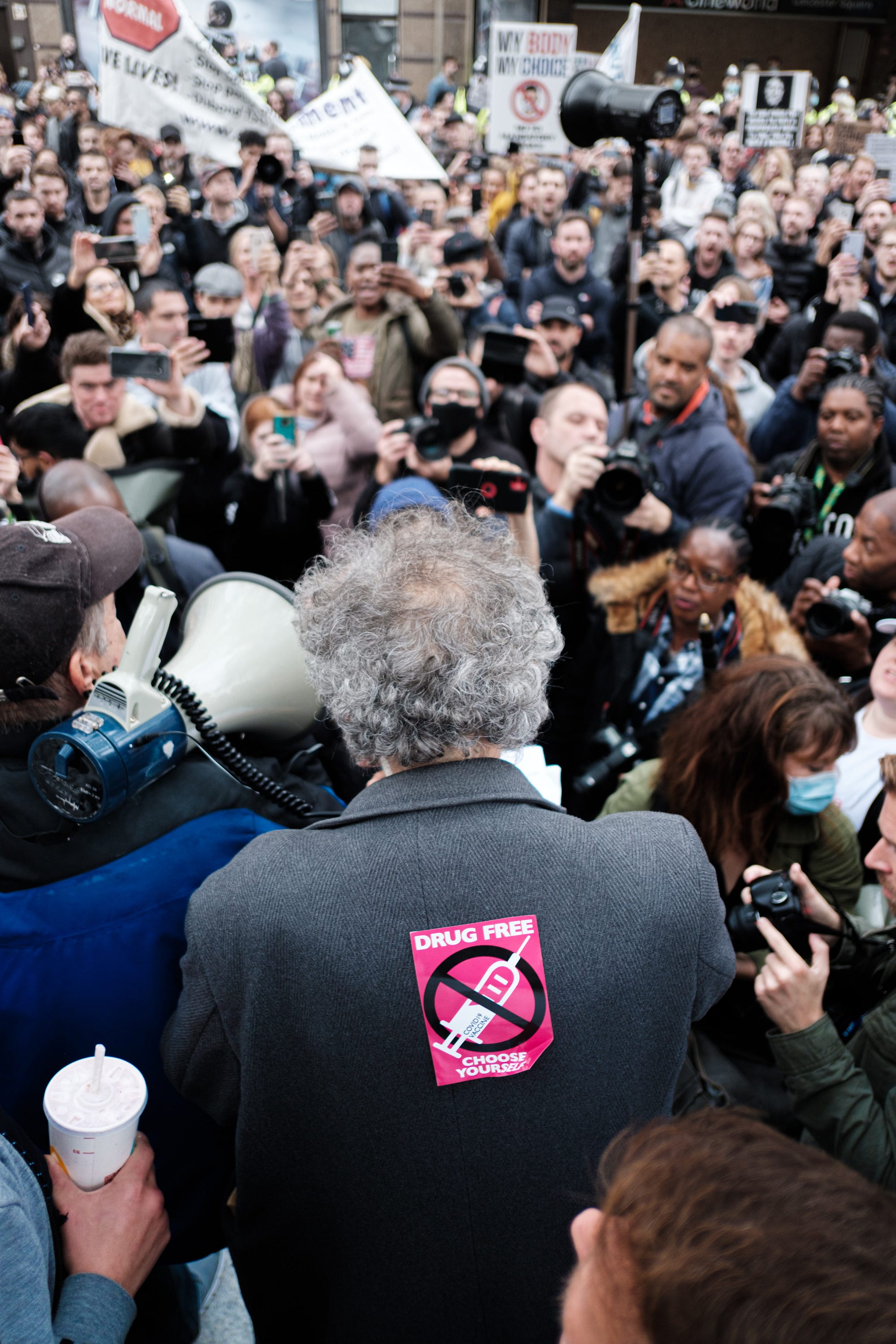 Man in front of a crowd, with his back to the camera, with an anti-vaccine sticker stuck to the back of his coat.