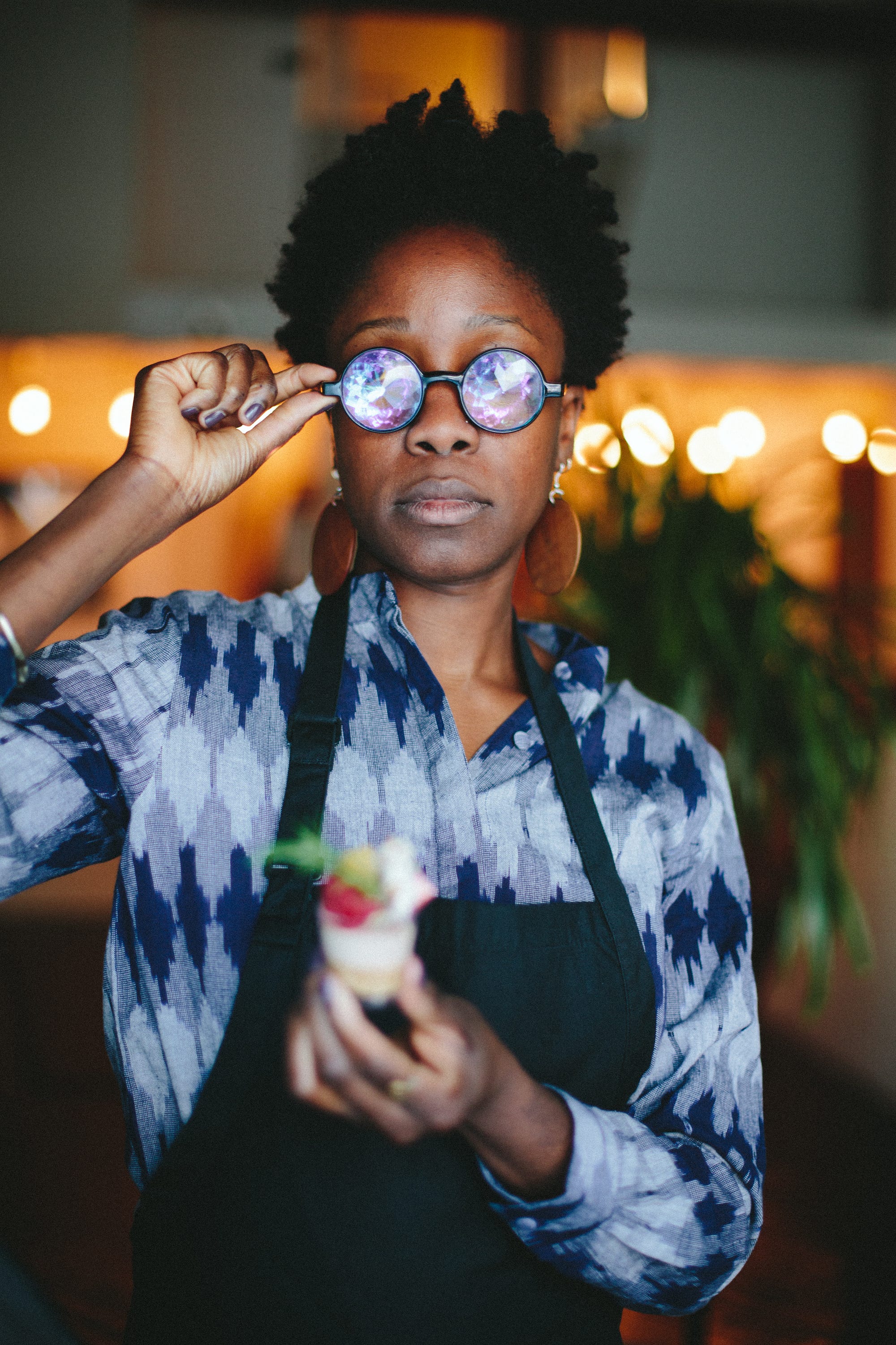 Yewande Komolafe wearing round glasses with iridescent lenses, holding a blurry dessert item.
