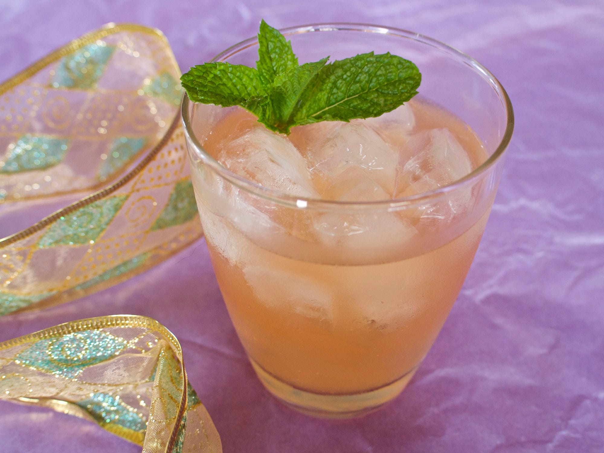 A cloudy, refreshing ginger-colored drink on ice, with a mint sprig and a lavender backdrop