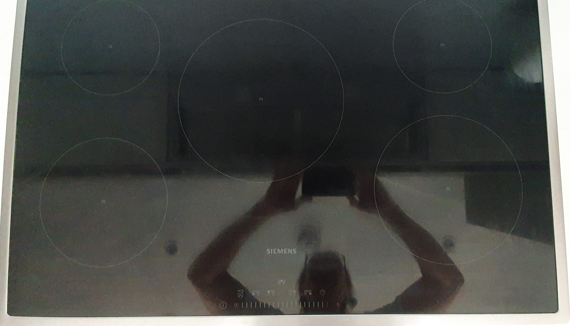 Overhead image of a induction hob. The surface is reflective black and shows the faint marking for five rings and the control dials to regulate the cooking rings.